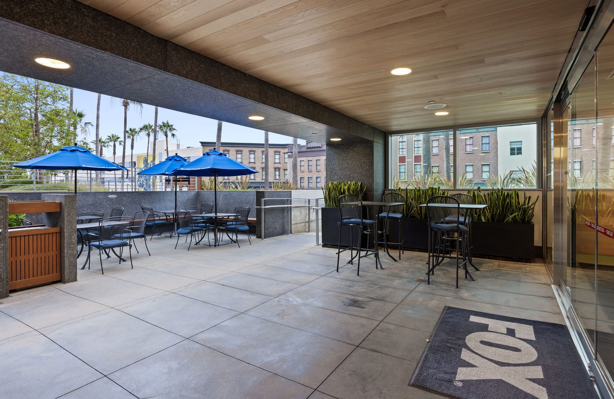 Outdoor patio with tables, chairs, blue umbrellas, a FOX doormat, and palm trees in the background.
