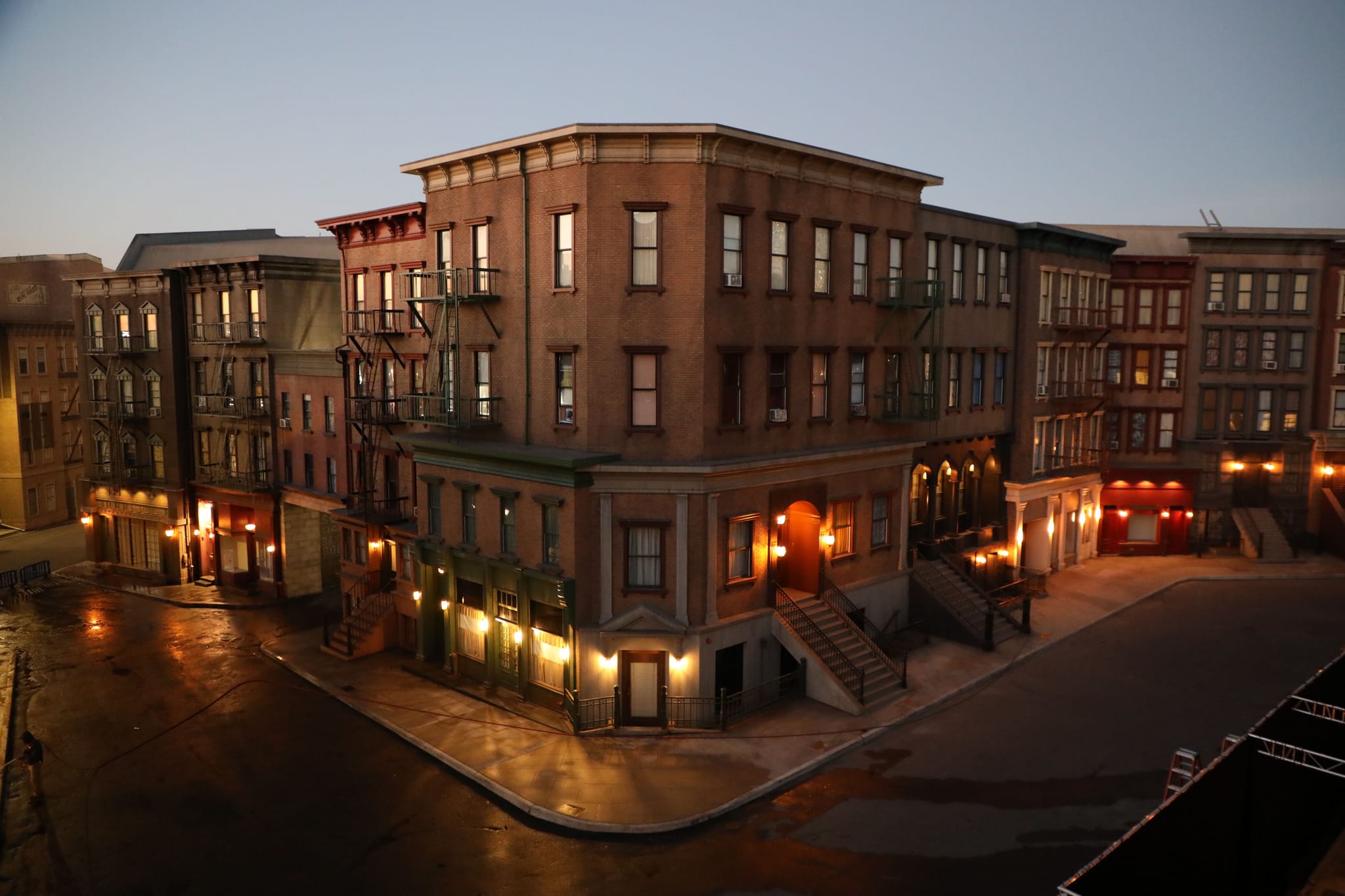 Multi-story brown buildings with fire escapes line a wet city street corner at twilight, their windows and streetlights casting warm reflections.