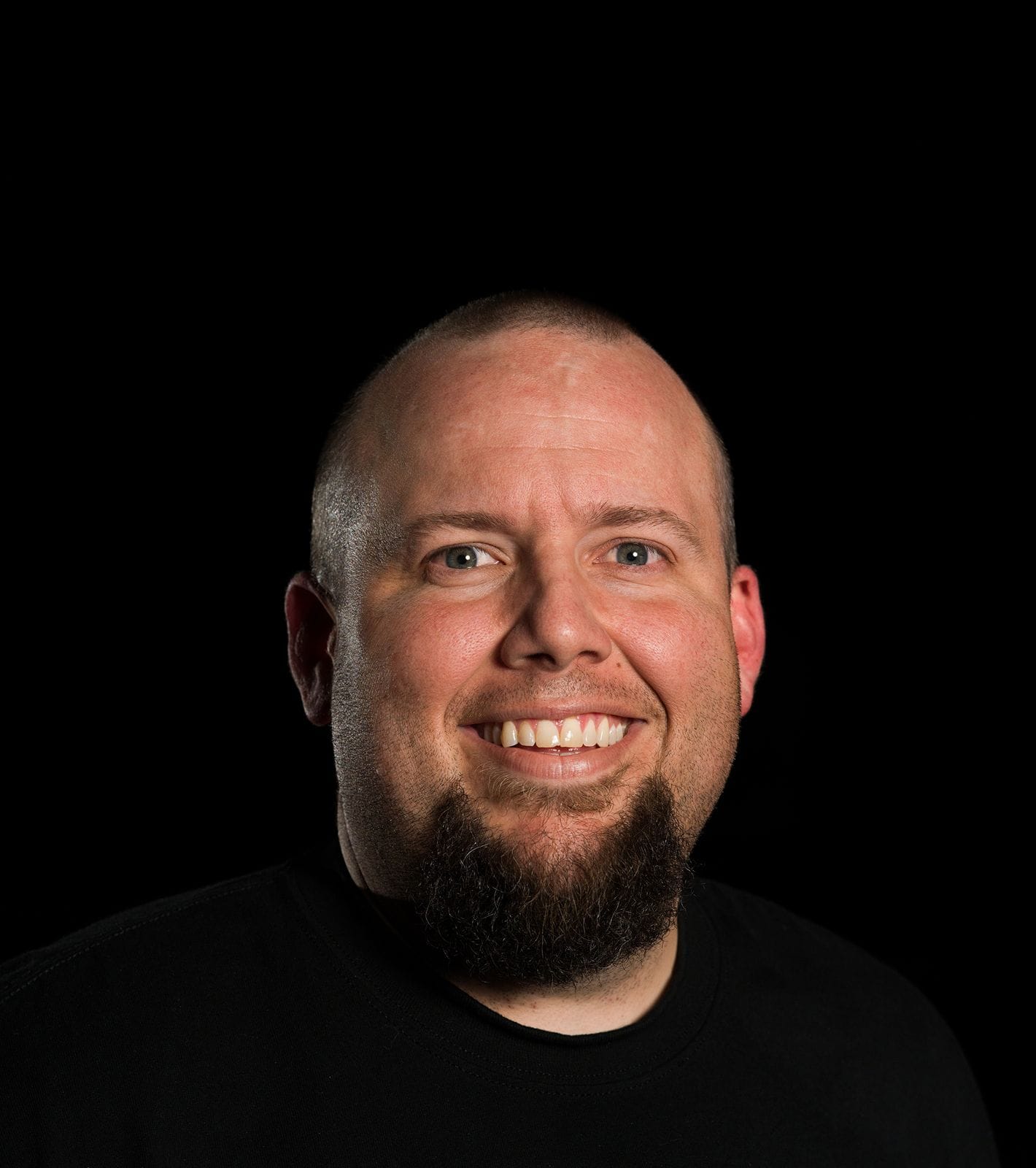 Smiling man with a shaved head and goatee in a black shirt against a black background.