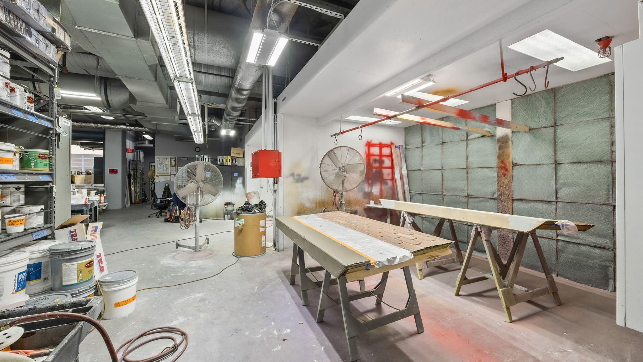 An industrial paint shop with shelves of paint, fans, and a spray booth with wooden planks on sawhorses.