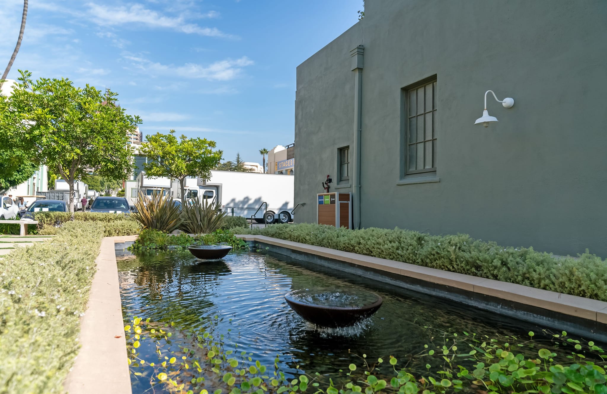 A long water feature with two bowl fountains surrounded by greenery, next to a pale green building on a sunny day.