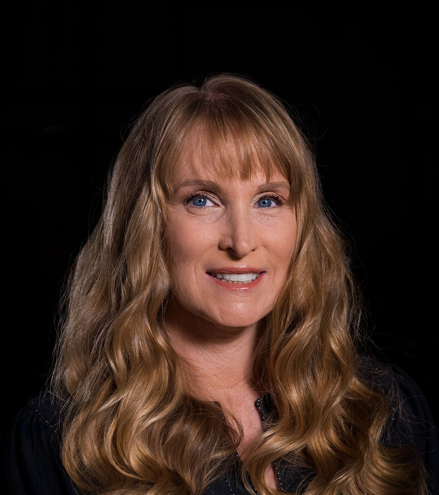 A smiling woman with long, wavy blonde hair and blue eyes against a dark background.