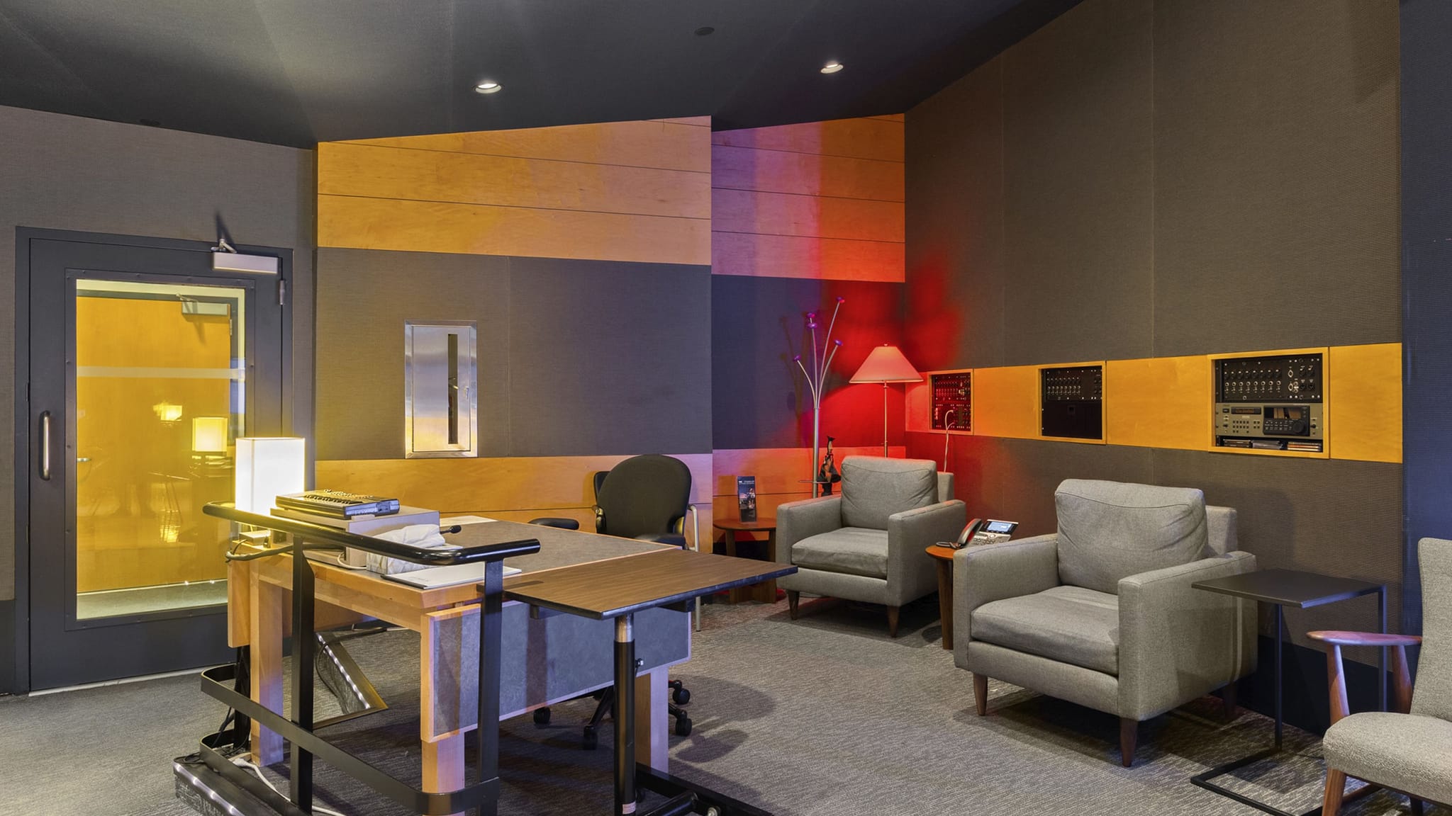 Modern lounge with a desk, two grey armchairs, sound-dampening walls, and red ambient lighting.