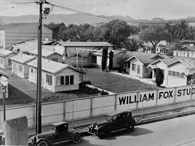 Black and white aerial view of William Fox Studios, showing rows of bungalows, green lawns, a fence with "WILLIAM FOX STUDIOS" painted on it, and vintage cars on a street.