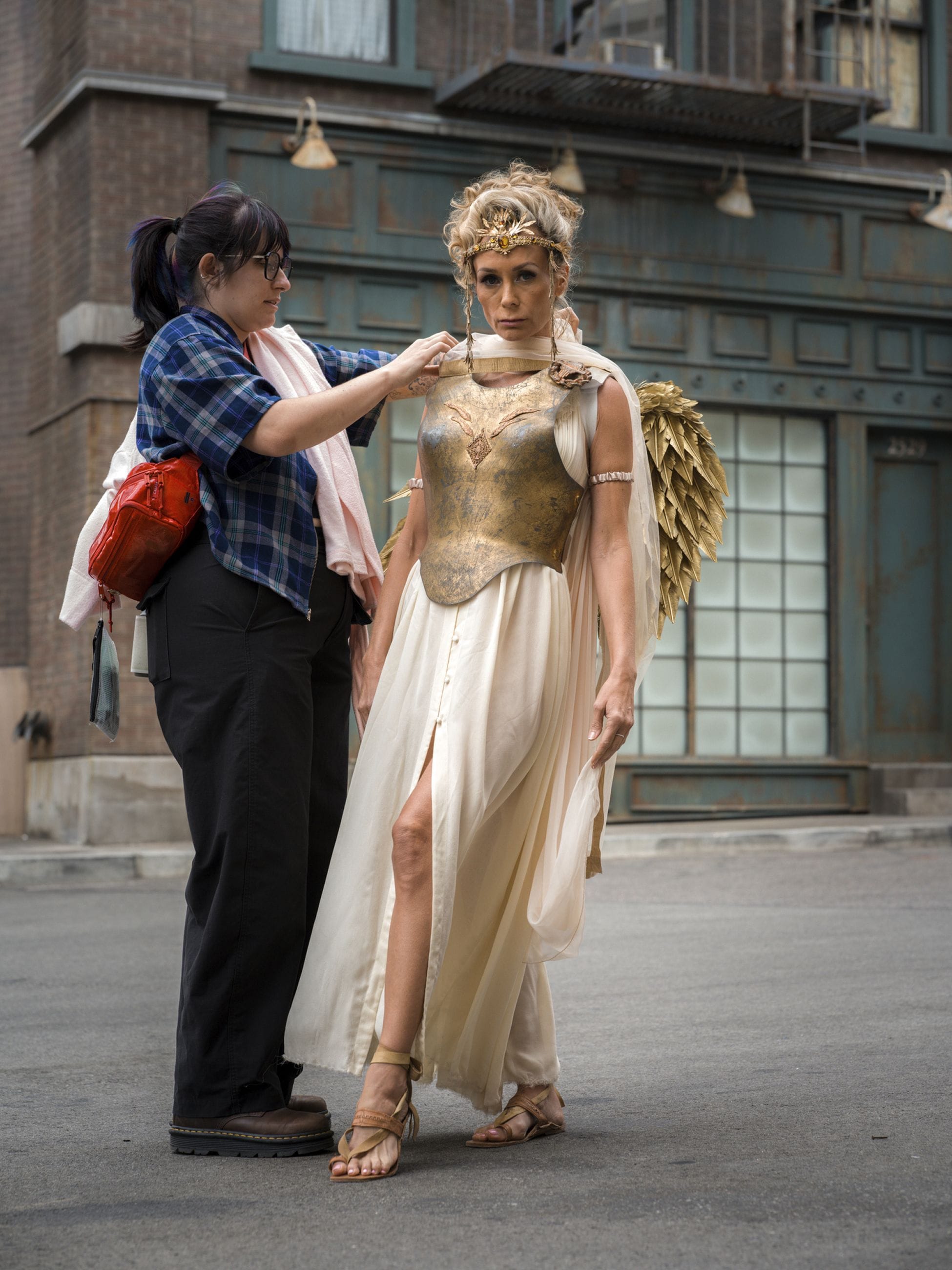 A costume designer adjusts a golden breastplate on an actress dressed as an angelic figure with golden wings and a crown.