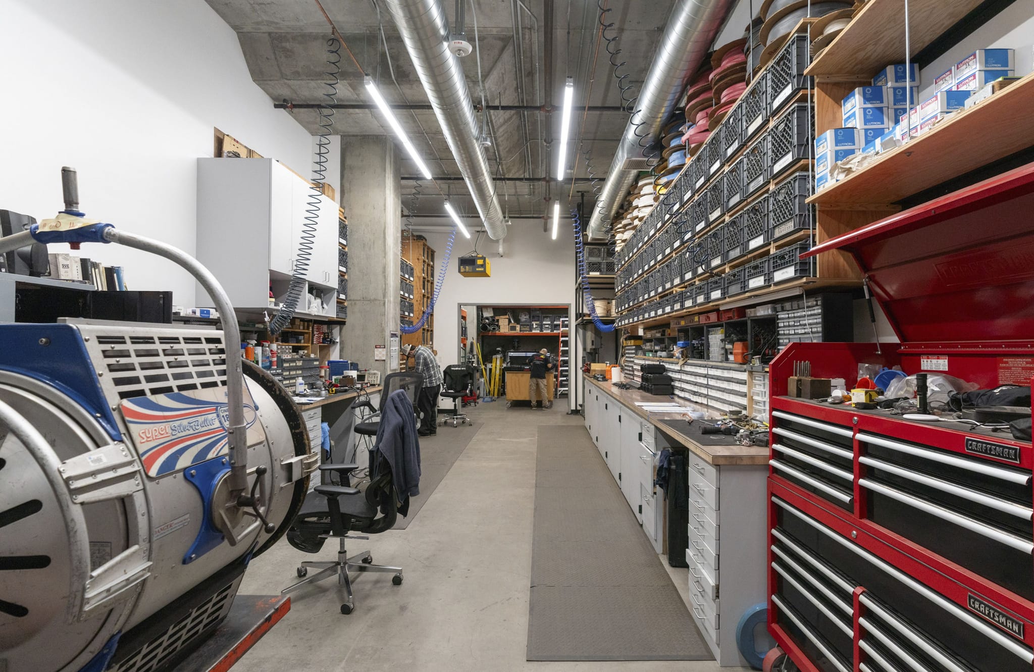 An organized workshop with a large industrial light or machine, workbenches, shelves filled with parts in crates, and a red tool chest.