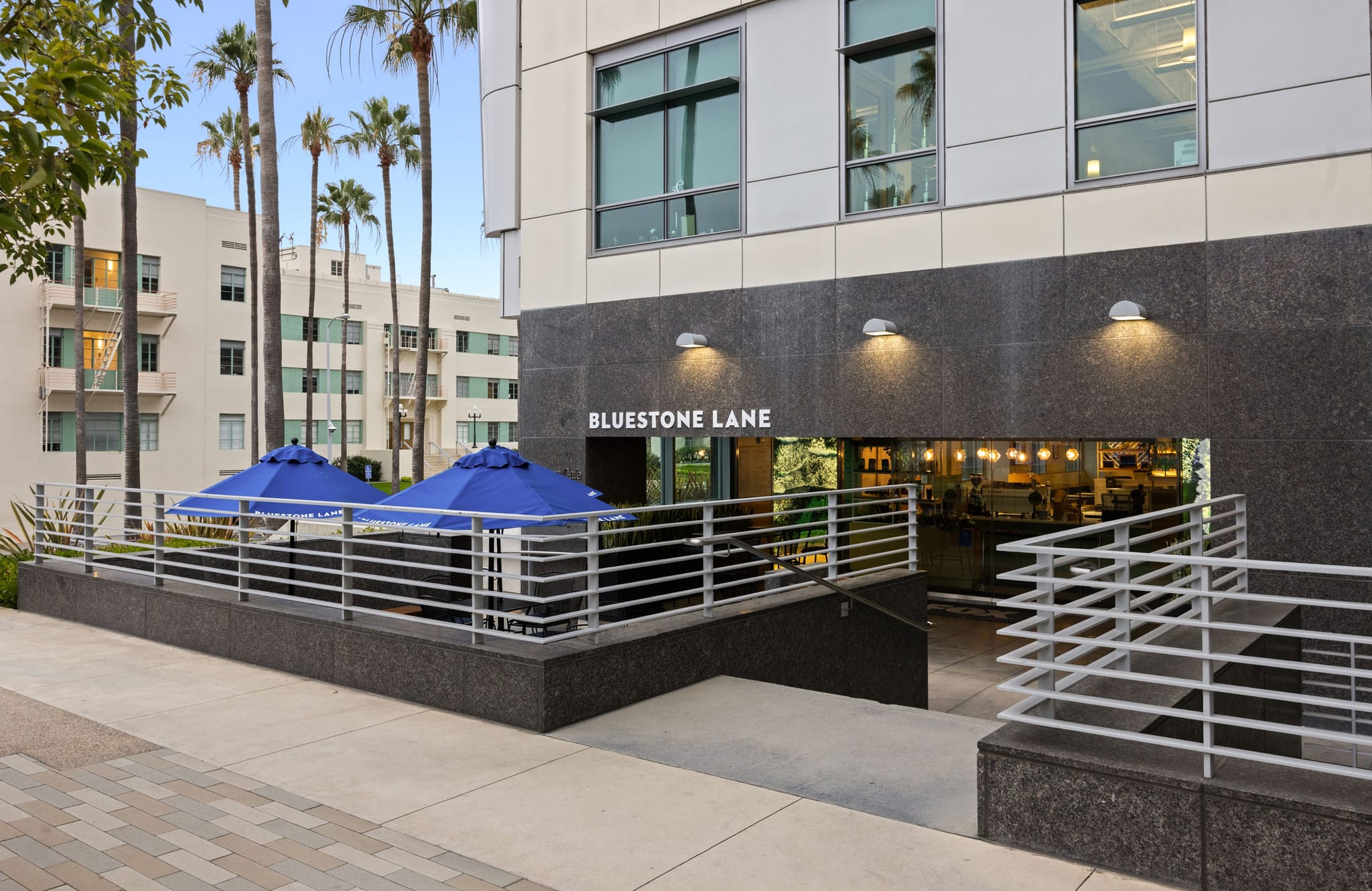 Exterior of Bluestone Lane cafe with outdoor seating under blue umbrellas and an illuminated entrance.
