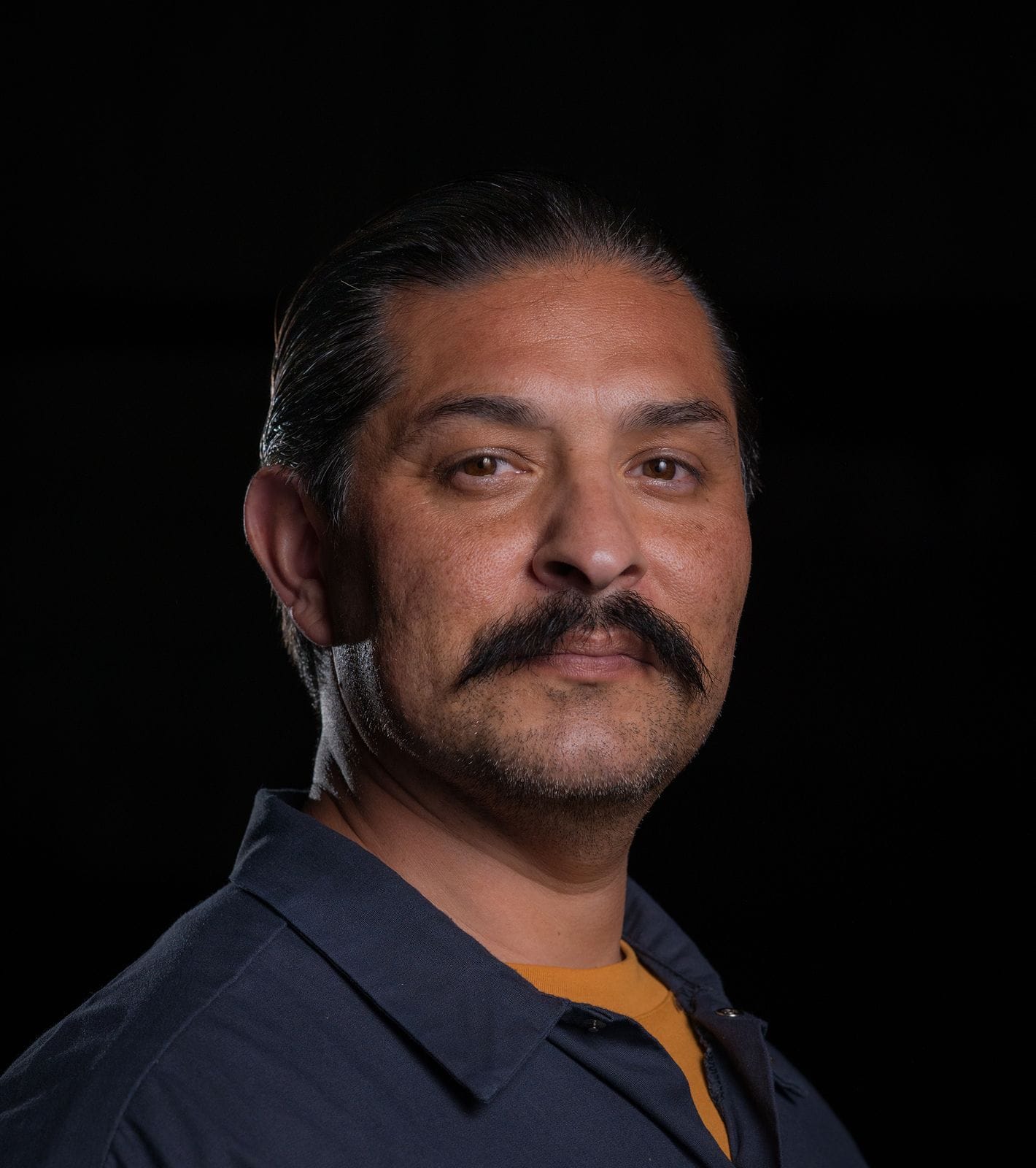 A man with dark slicked-back hair and a prominent mustache wears a dark blue collared shirt and looks at the camera.