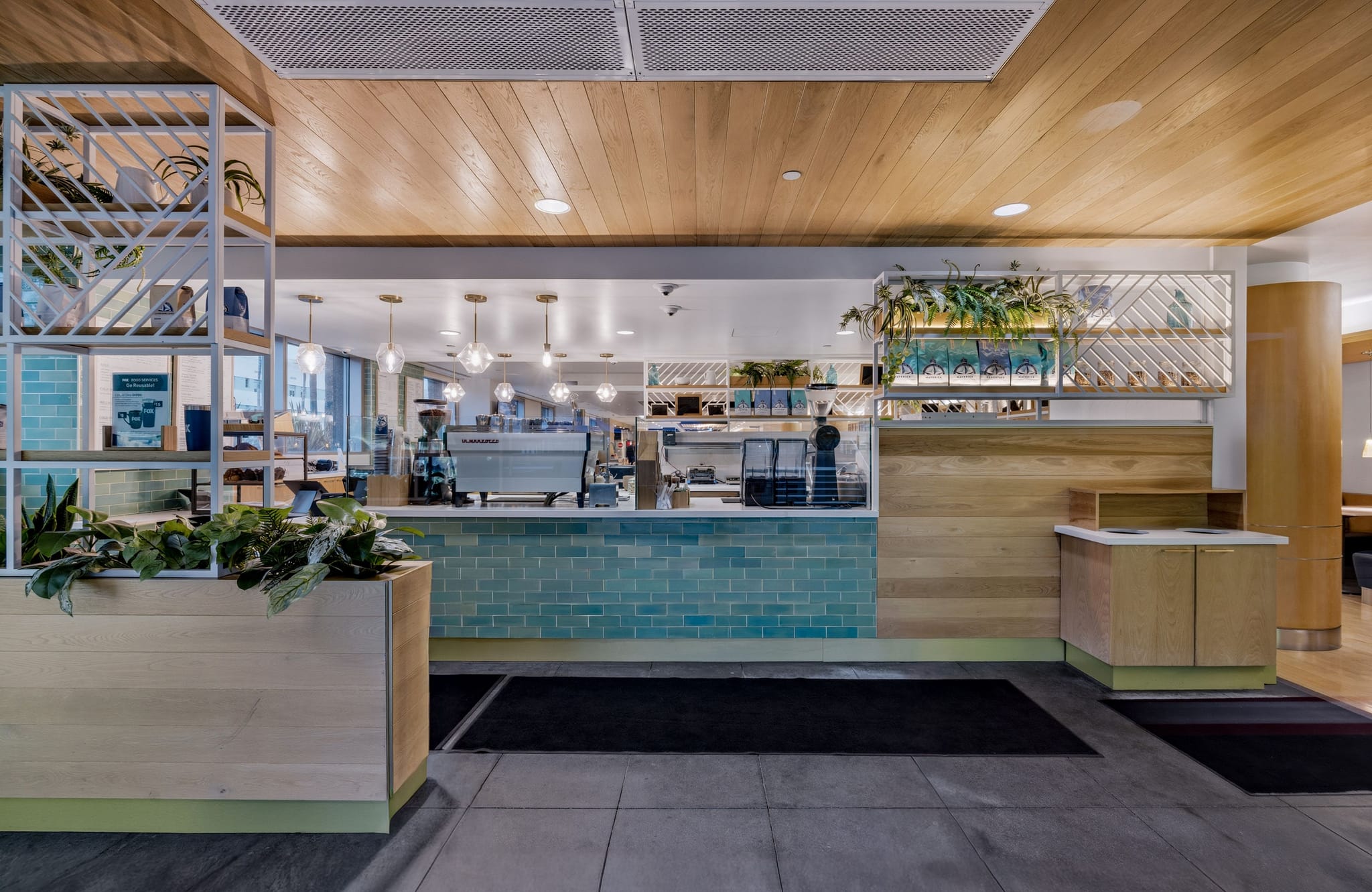A modern cafe counter with a large espresso machine, teal tile front, wooden walls and ceiling, and potted plants.