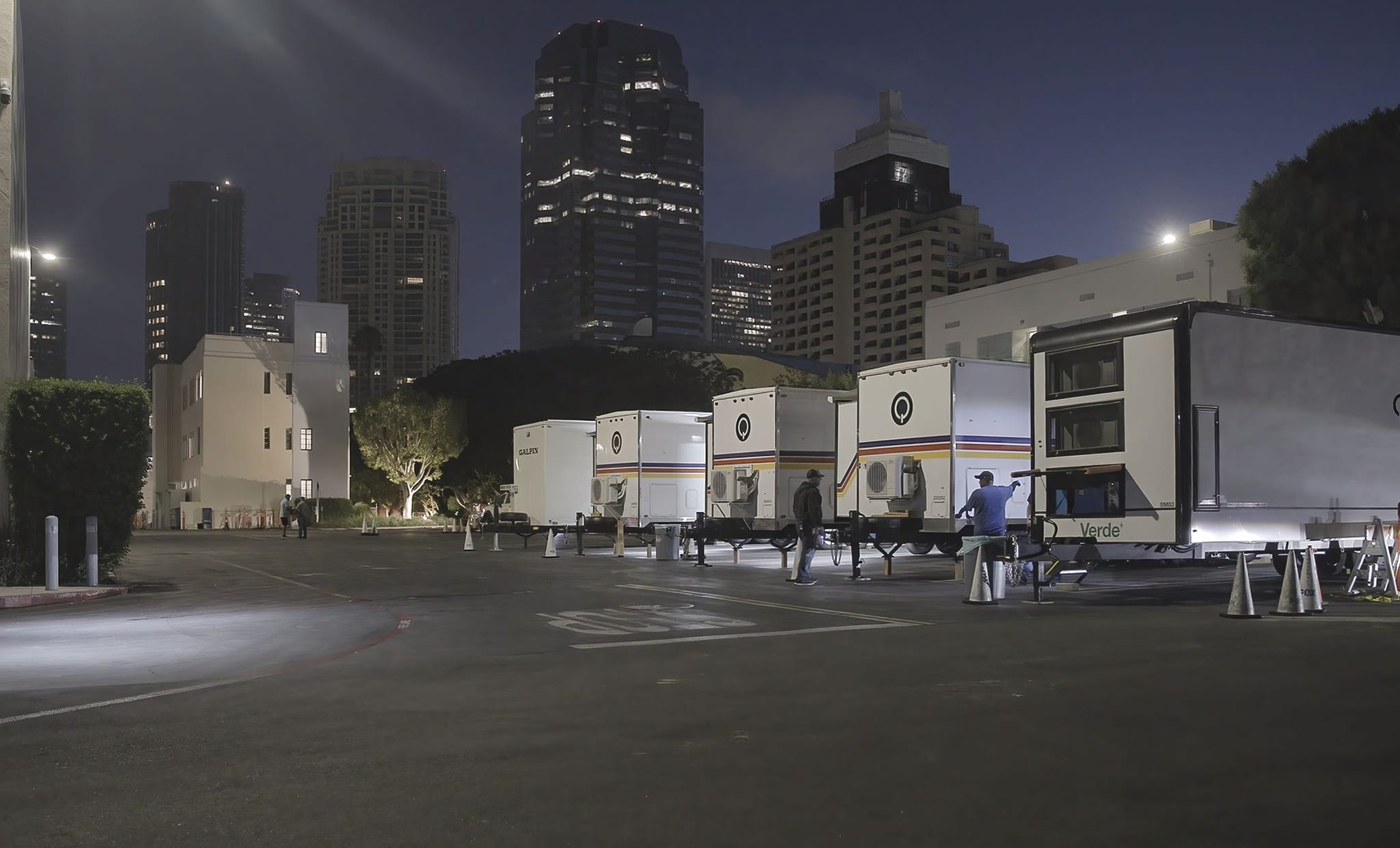 Night view of several white mobile units and people in a city parking lot, with illuminated skyscrapers in the background.