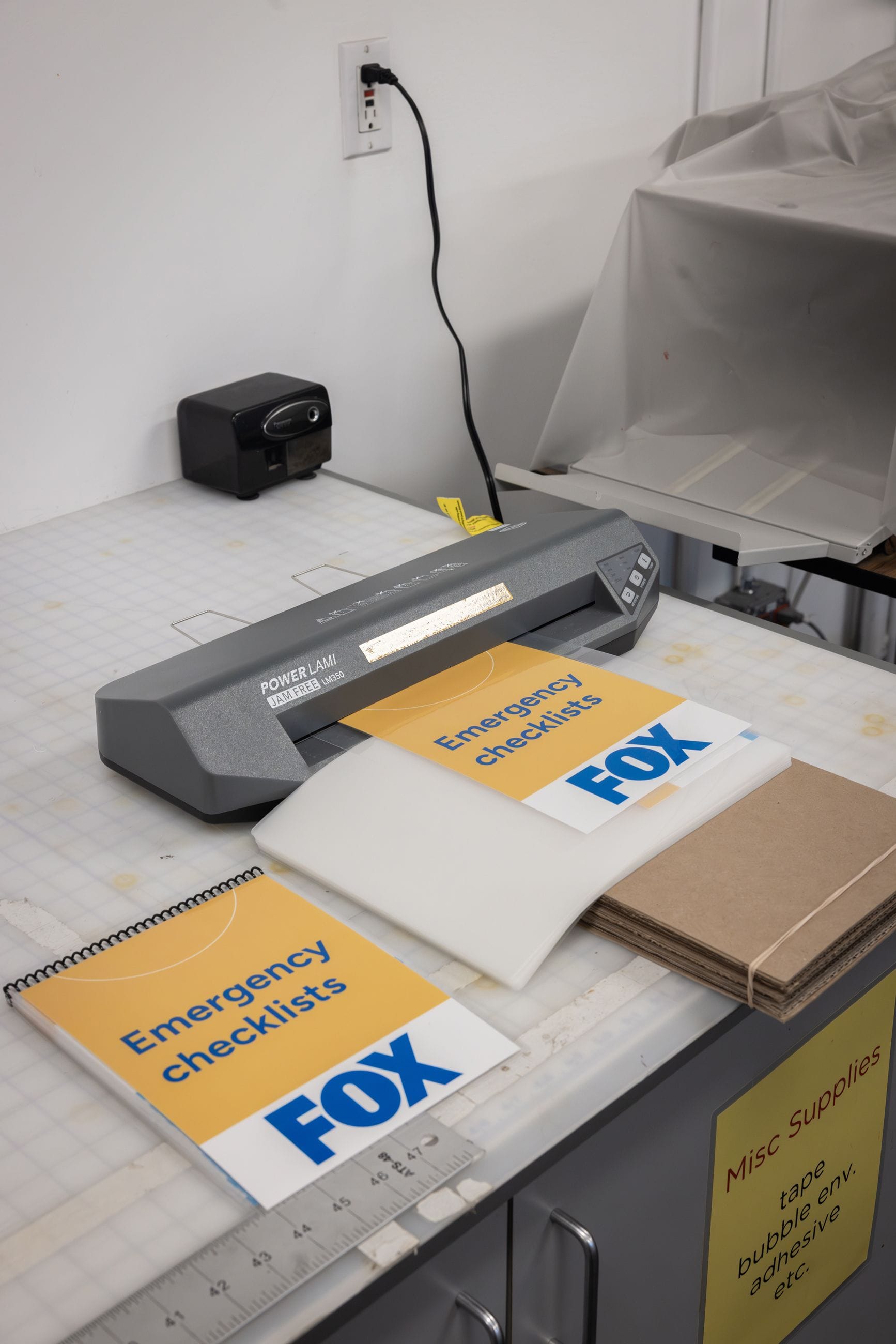A grey laminator processes a yellow page labeled "Emergency checklists FOX," while a finished, spiral-bound booklet with the same title sits nearby on a white workspace.