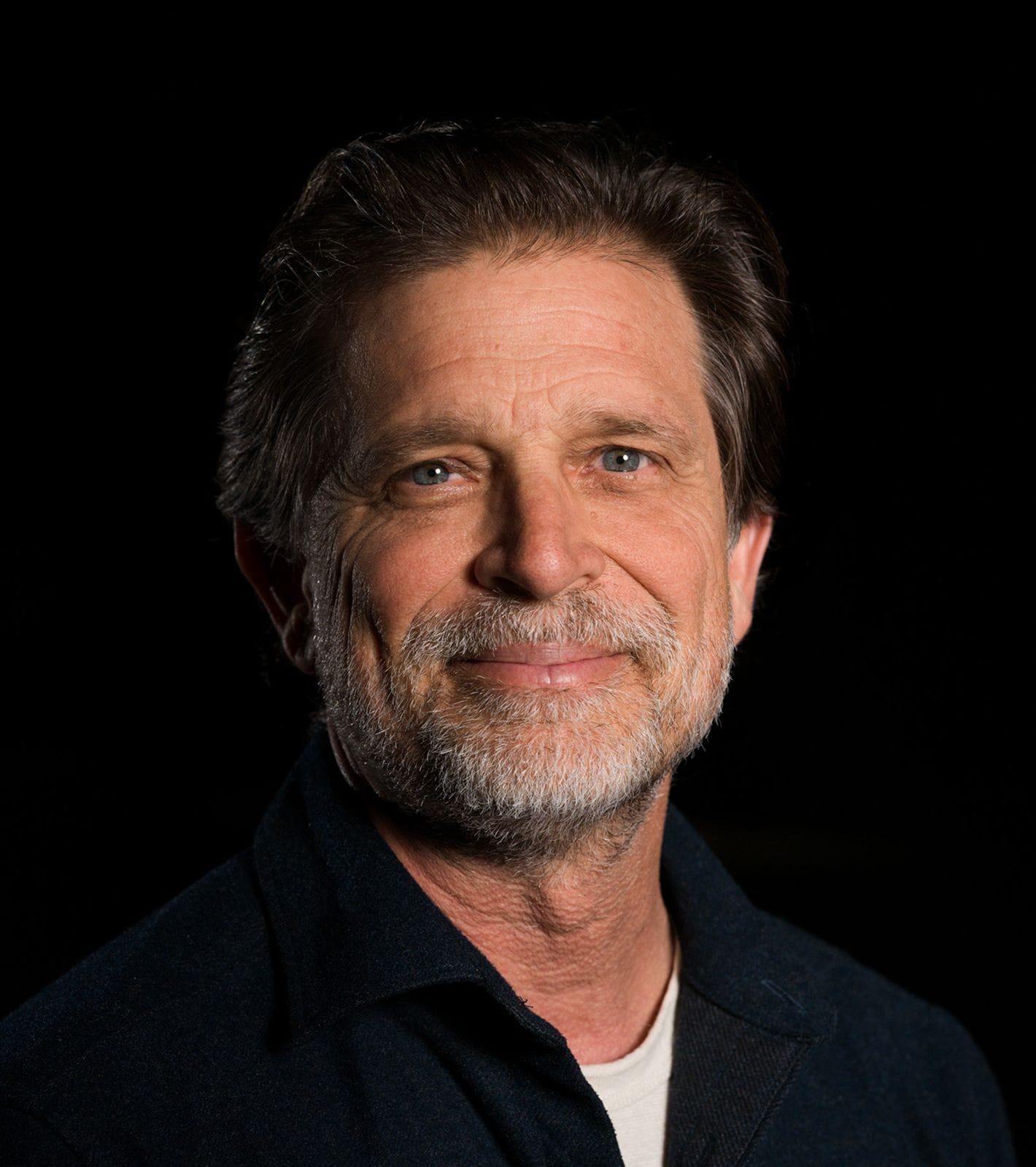 Headshot of a smiling man with a graying beard and brown hair against a dark background.