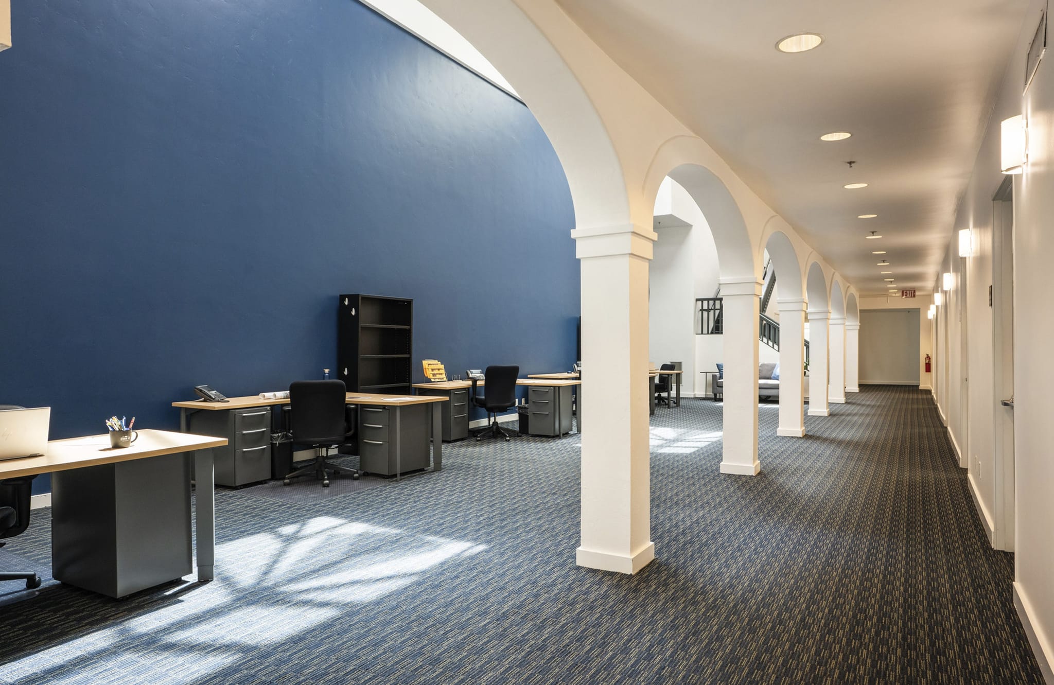 Open office space with a blue wall, multiple desks, a blue patterned carpet, and a hallway lined with white arched pillars.