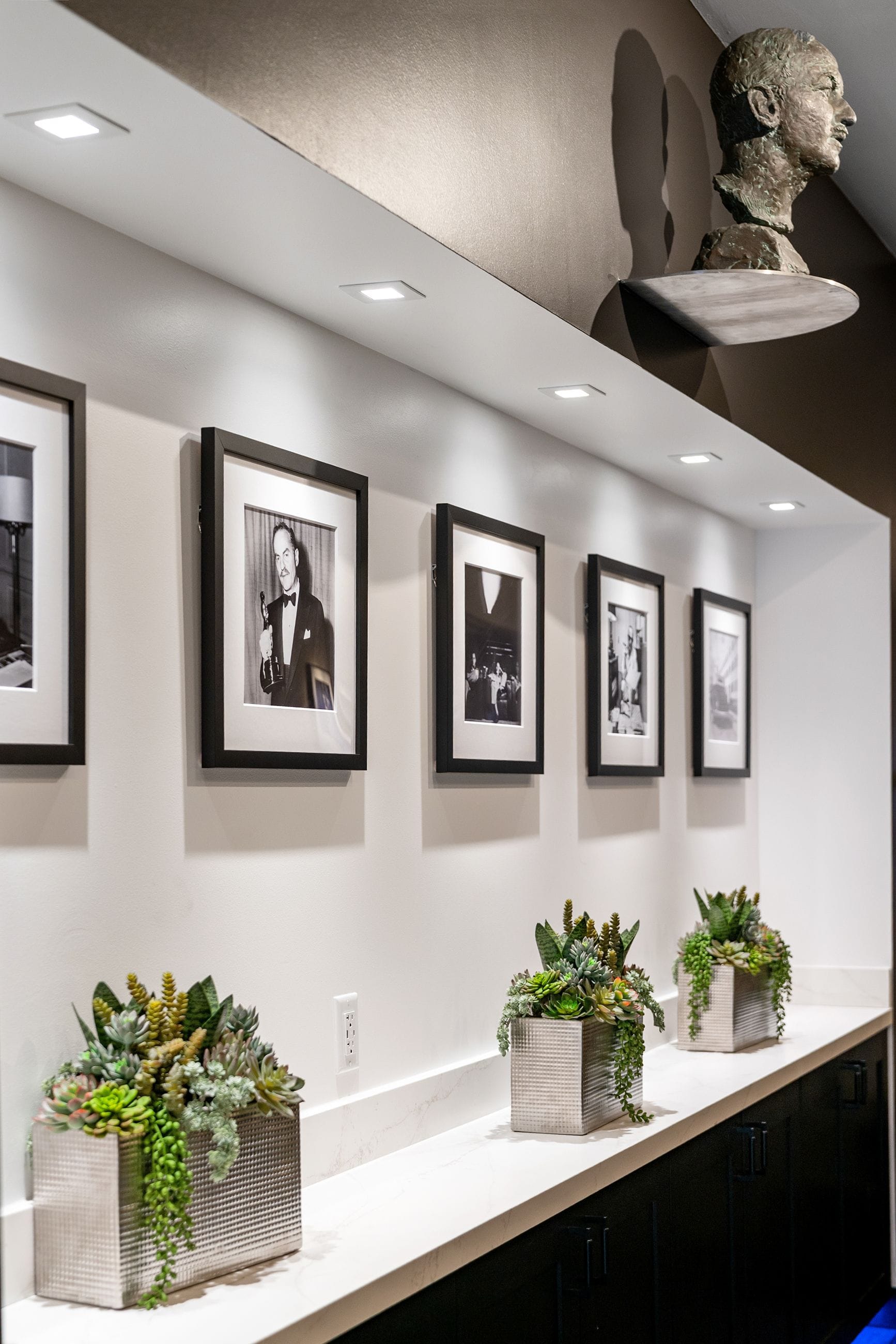 A modern hallway with framed black and white photos, succulent planters on a white counter, and a bronze bust on a shelf.