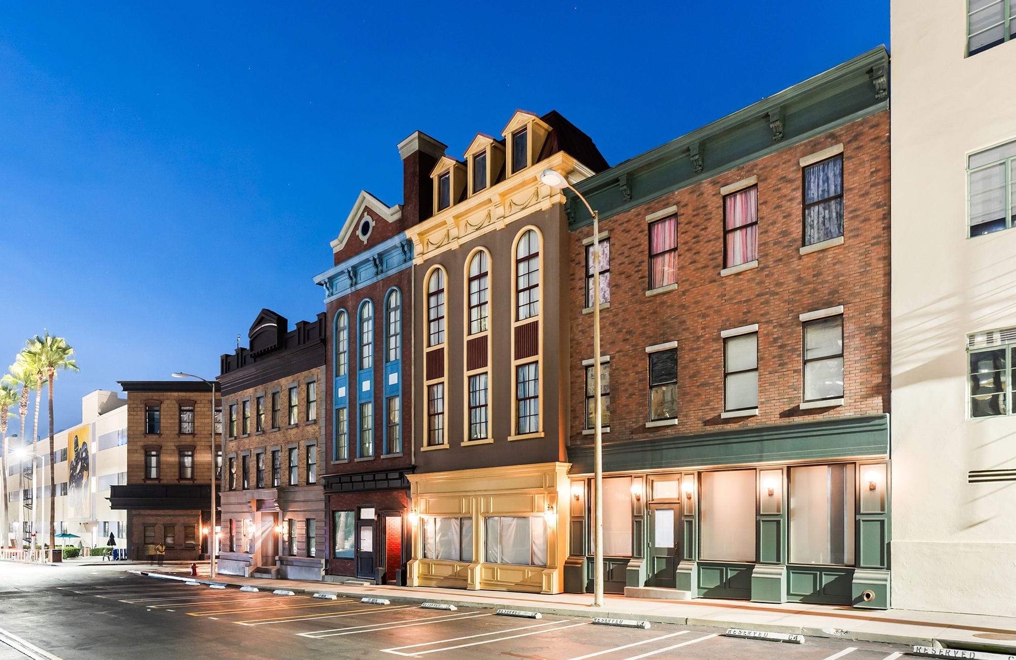 A street lined with diverse buildings and storefronts at dusk, under a clear blue sky, with palm trees on the left.
