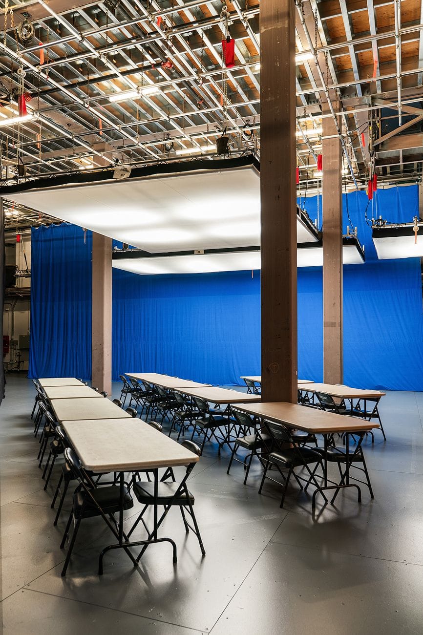 An empty studio setup with rows of folding tables and chairs, a blue backdrop, and overhead lighting equipment.