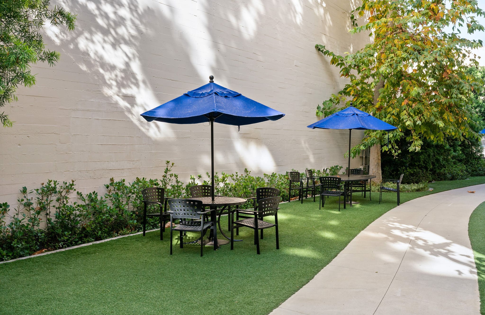 Outdoor patio area with several tables, chairs, and blue umbrellas on artificial turf, next to a curved path and a light-colored wall with foliage.