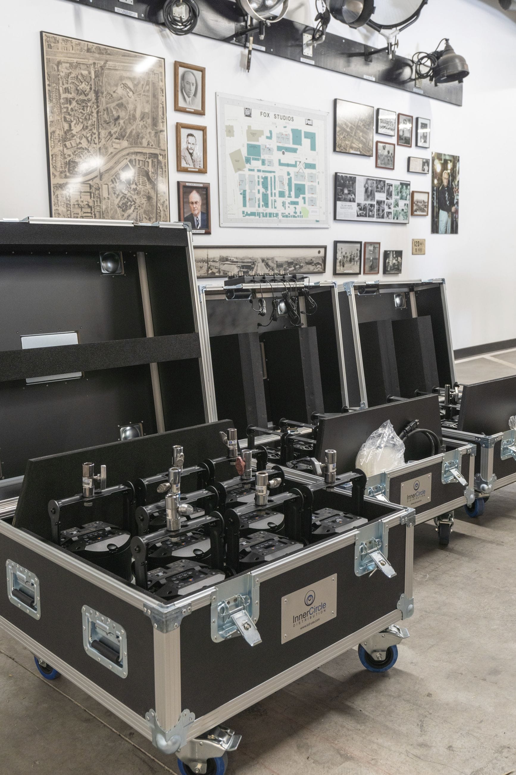 Open road cases filled with stage lighting fixtures, with framed studio memorabilia on a wall in the background.
