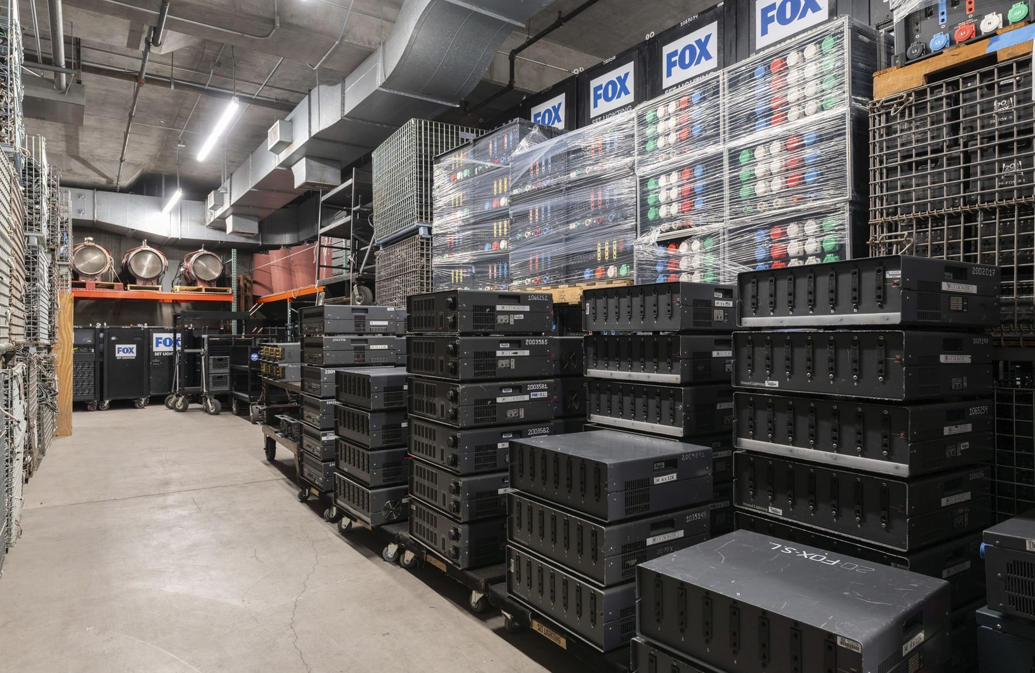 A storage room filled with stacks of black electronic equipment, stage lights, and FOX-branded boxes.