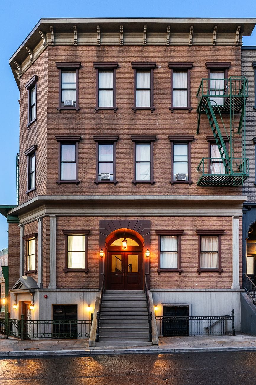 A multi-story brick building with a central entrance staircase, many windows, and a green fire escape, under a twilight sky.