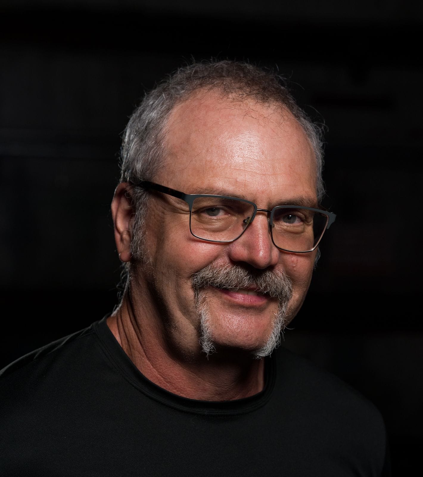 A man with graying hair, glasses, a mustache, and a goatee smiles against a dark background.