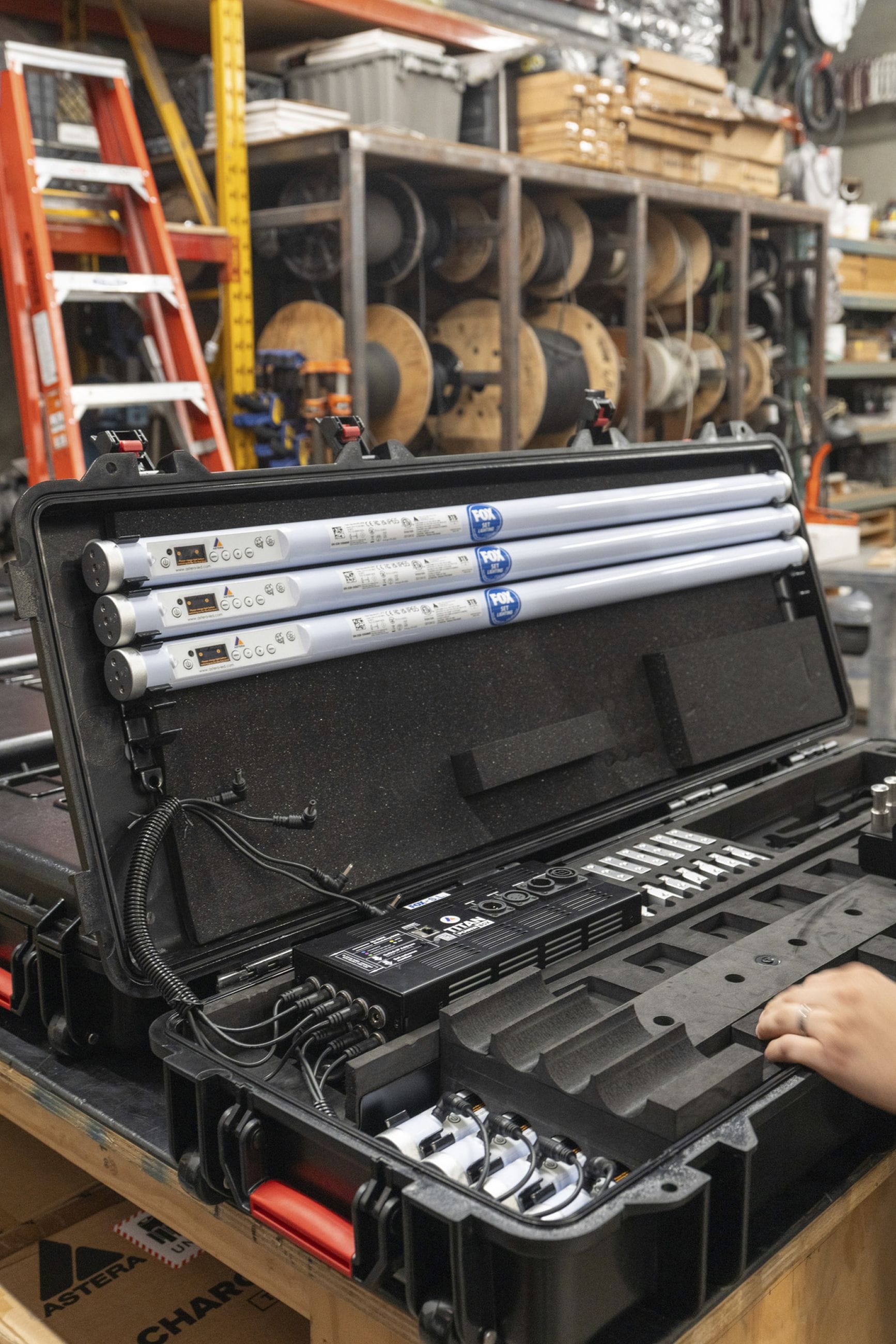 An open black hard case reveals three professional tube lights, a charger, and cables, in a warehouse setting.