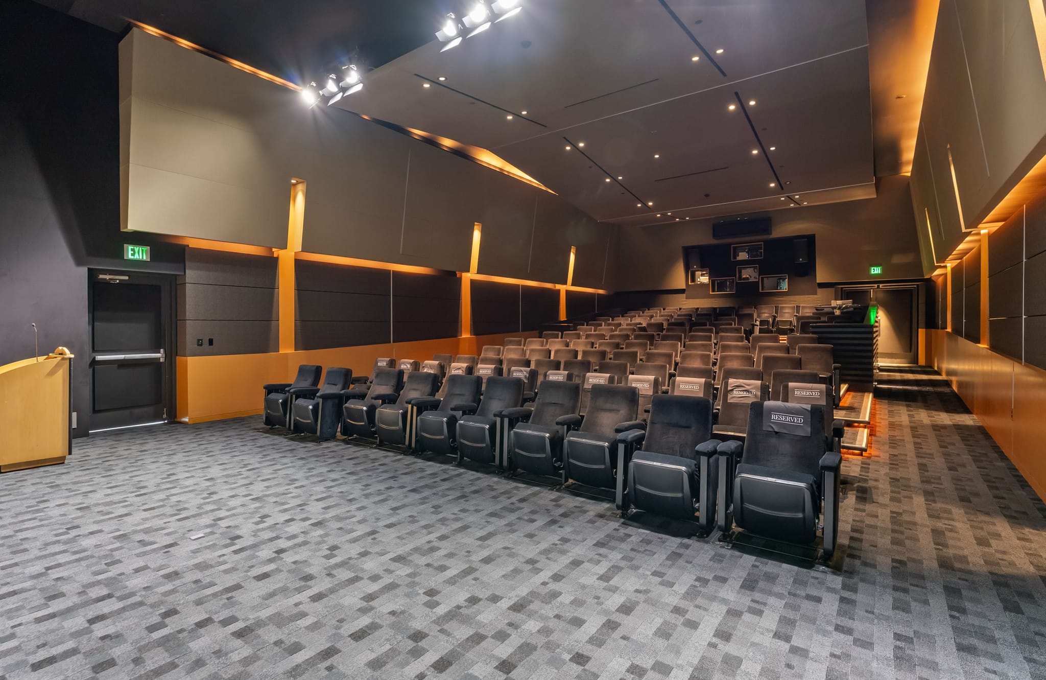 Empty modern auditorium with rows of dark seats, gray patterned carpet, a wooden podium, and dark walls with orange accents.