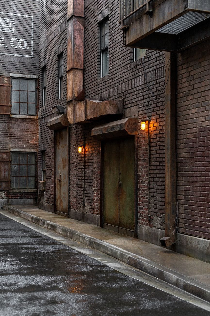 A gritty, wet urban alley lined with dark brick buildings, rusty doors, and glowing industrial lights.