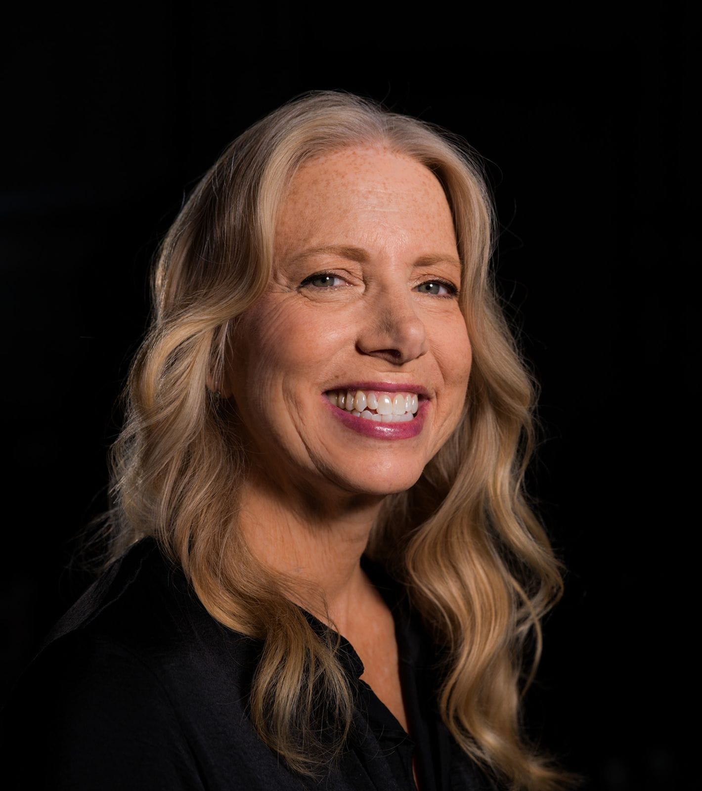 A smiling woman with long wavy blonde hair and a black shirt against a dark background.