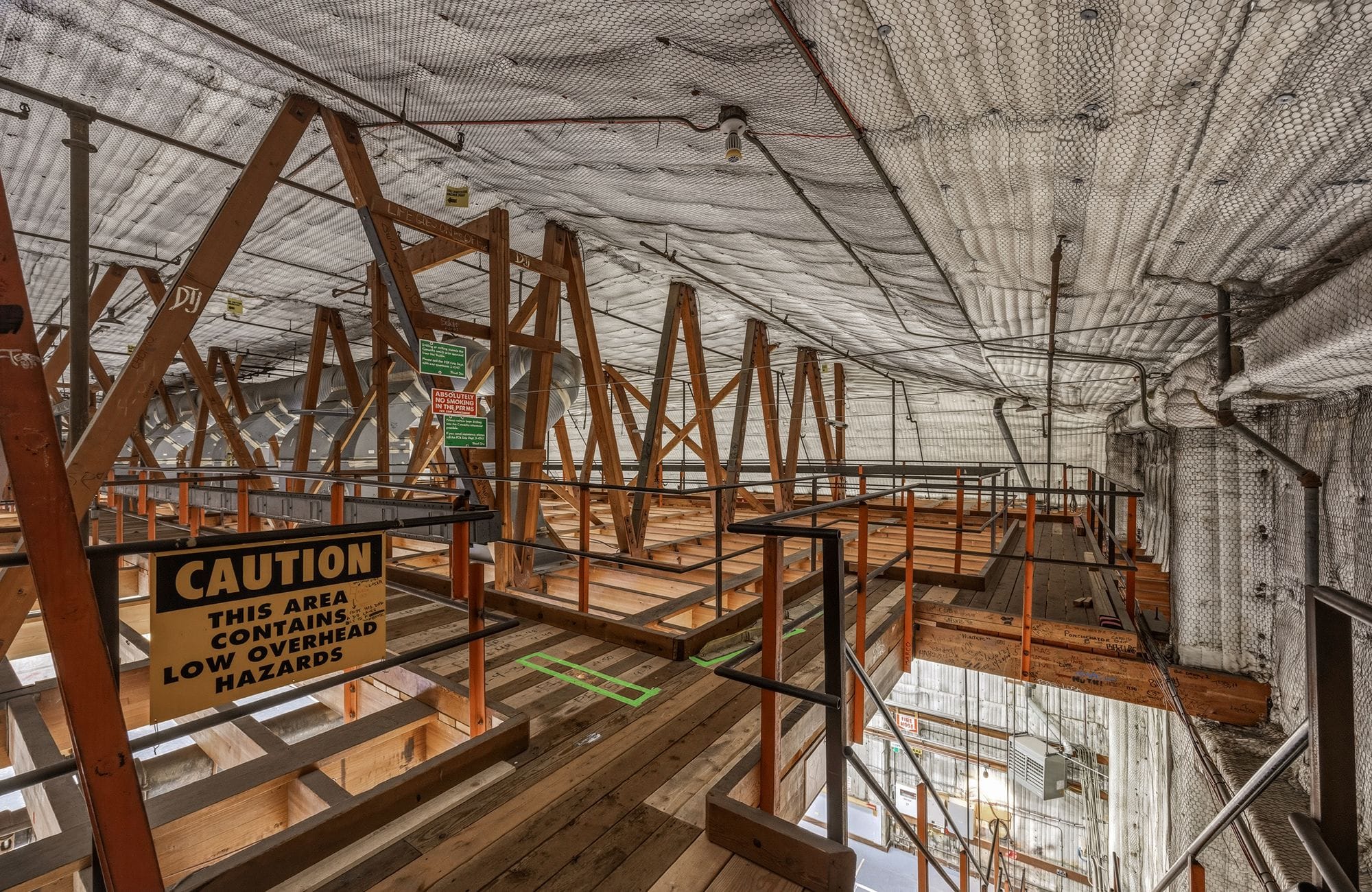 Industrial interior with exposed wooden beams, platforms, and a 'CAUTION LOW OVERHEAD HAZARDS' sign.