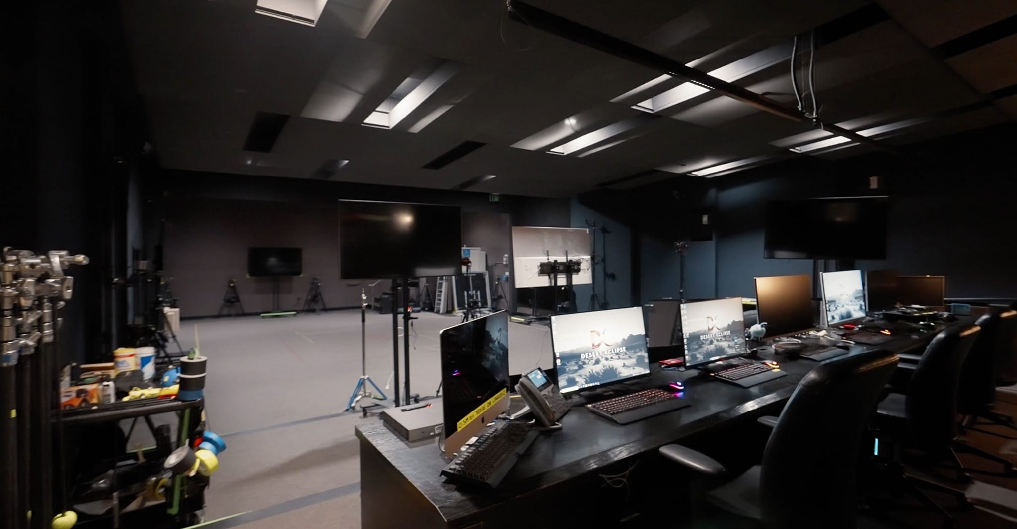 A man wearing a motion capture suit and a helmet with a facial camera is assisted in a studio, with a virtual environment displayed on a large screen.