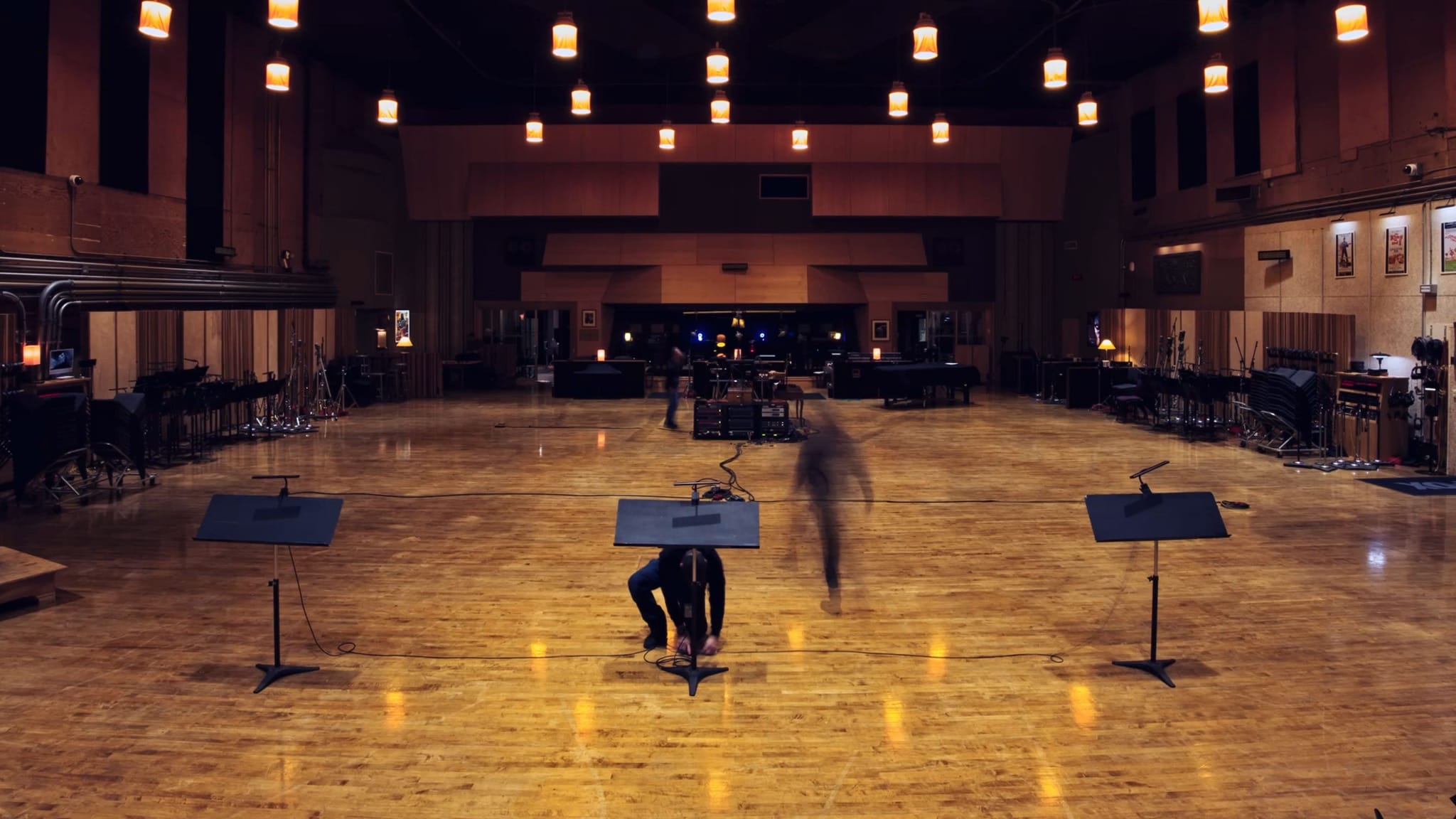 A large, warm-lit recording studio with two music stands in the foreground and two figures, one blurred by motion.