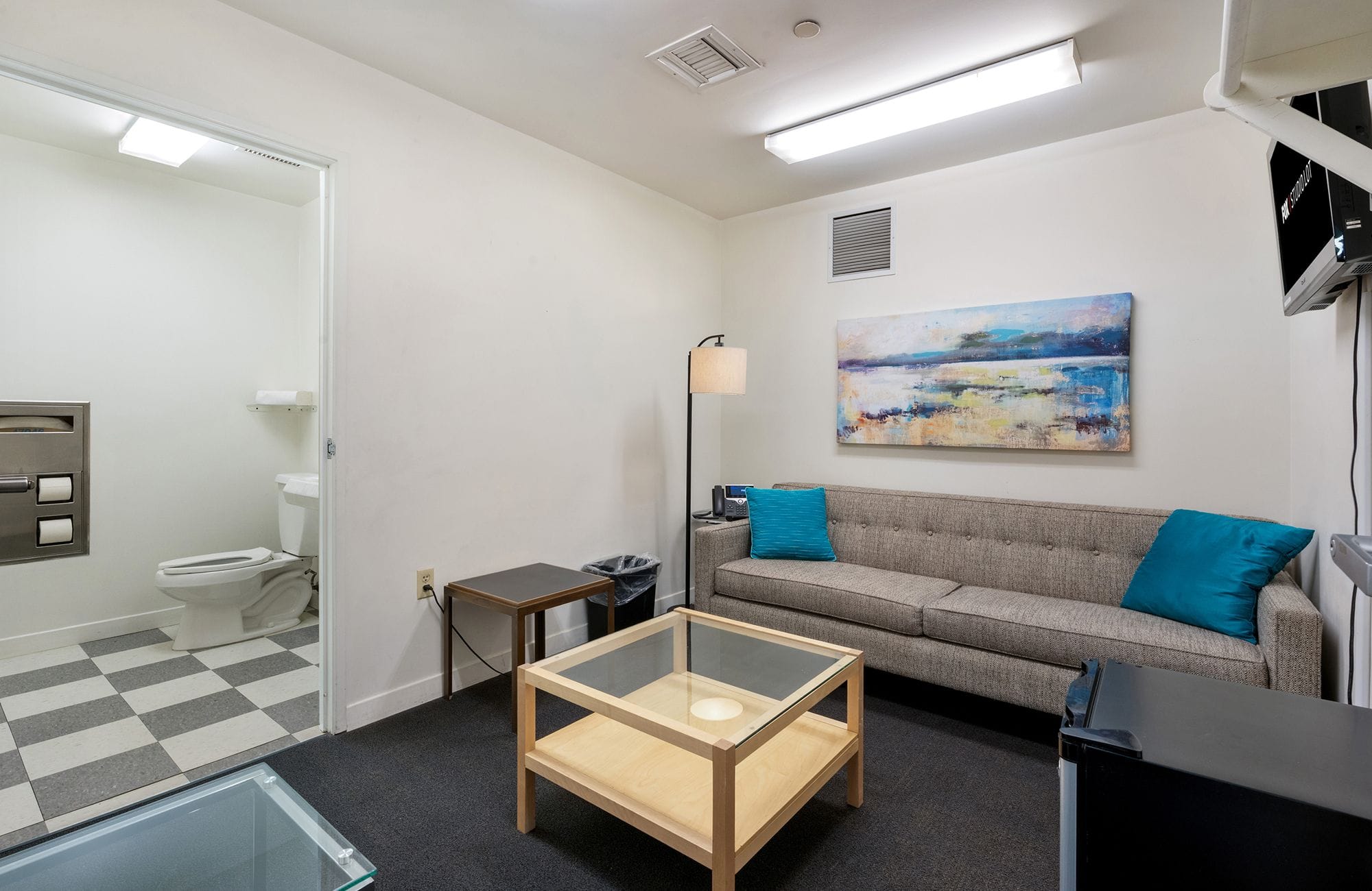 A modern room with a grey sofa, abstract art, wood and glass tables, and an open doorway to a bathroom with a checkered floor.