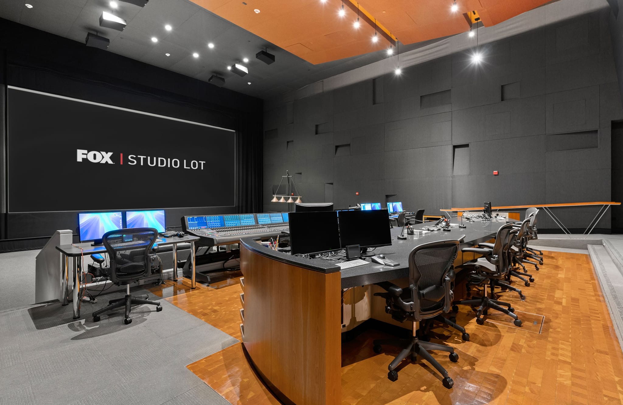 Modern control room with a large screen displaying "FOX | STUDIO LOT" above a console with multiple monitors and office chairs.
