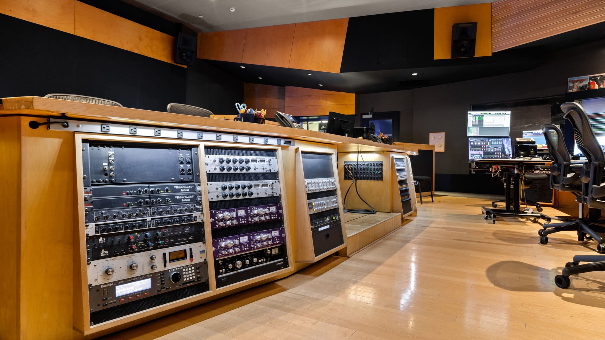Professional audio studio control room with a long wooden console containing numerous racks of audio equipment, light wood floors, and computer workstations.