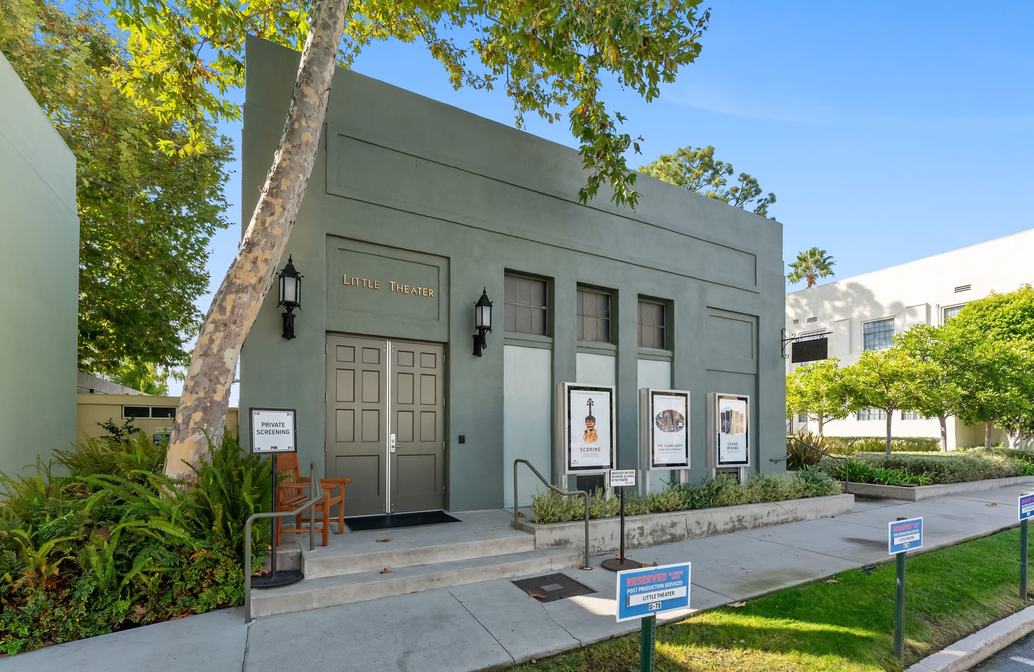 A muted green building with "LITTLE THEATER" above double doors, surrounded by trees and a paved walkway on a sunny day.