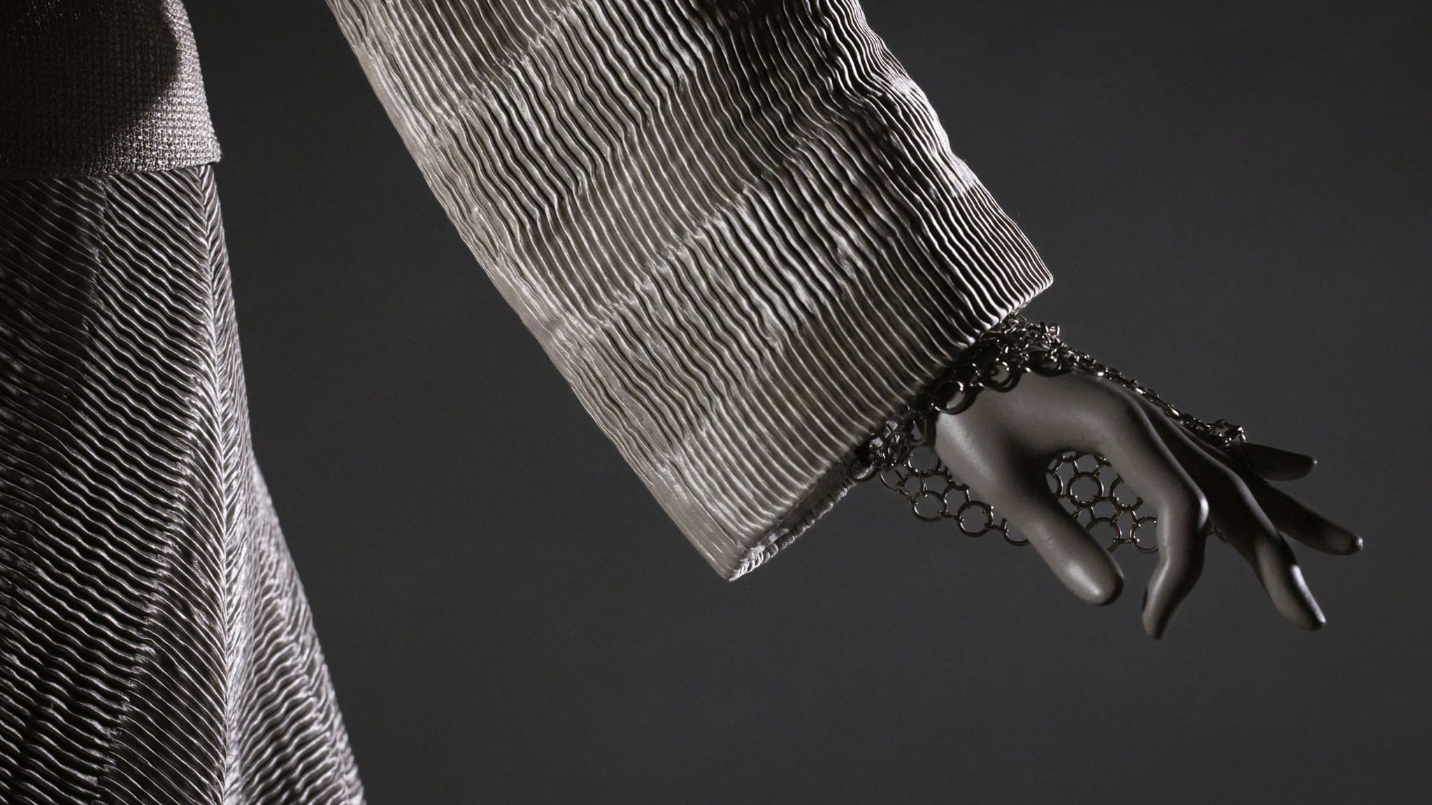 Mannequin arm in a grey wavy-textured garment with a metallic hand harness connecting a bracelet to rings.