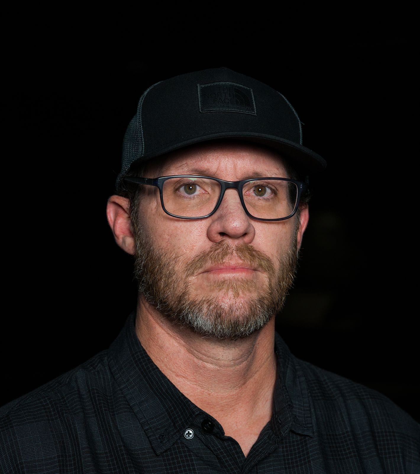 Man with beard, glasses, and black baseball cap wearing a dark plaid shirt against a black background.
