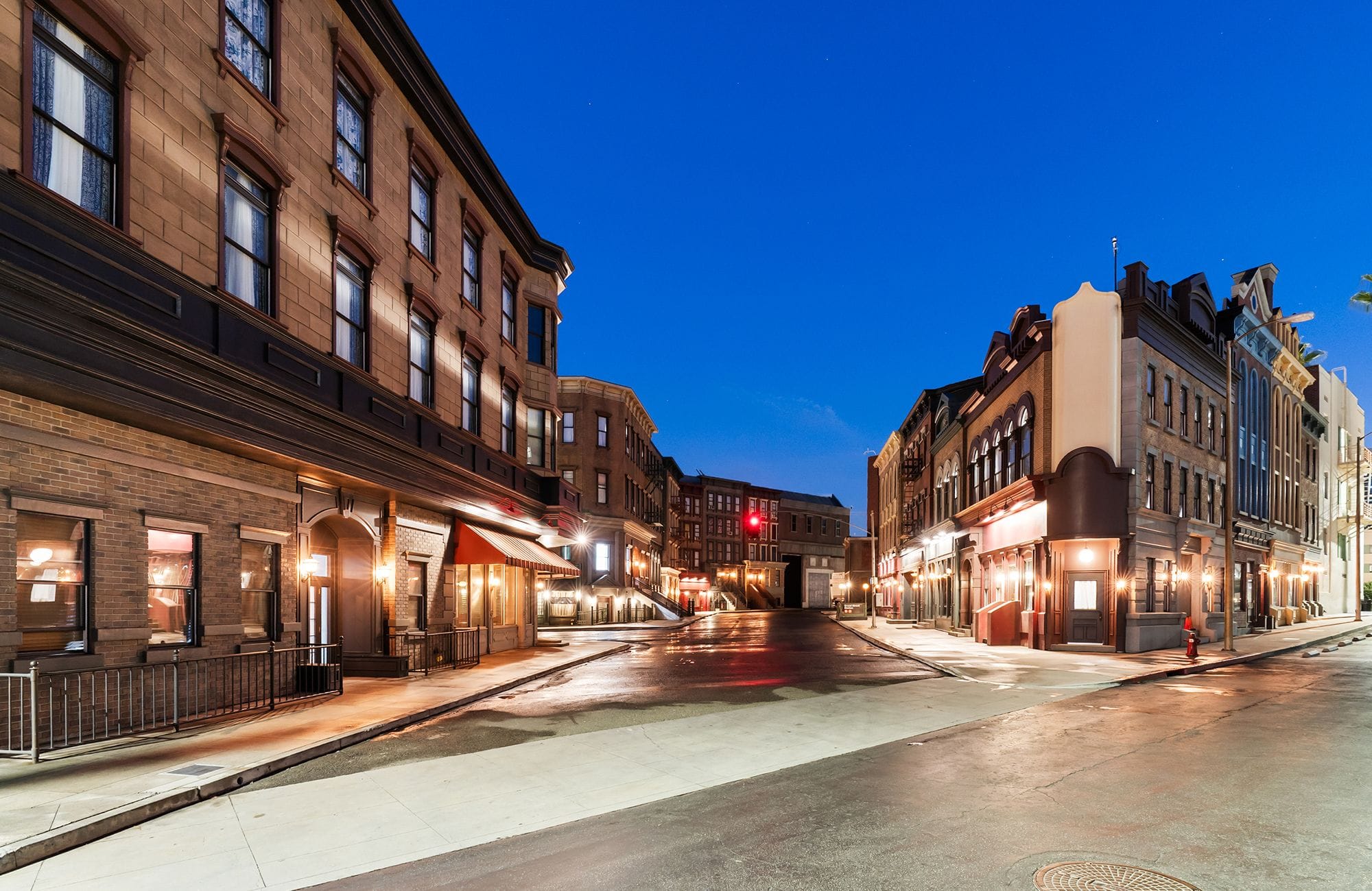 A historic city street at twilight, with wet pavement reflecting glowing storefronts and building lights.