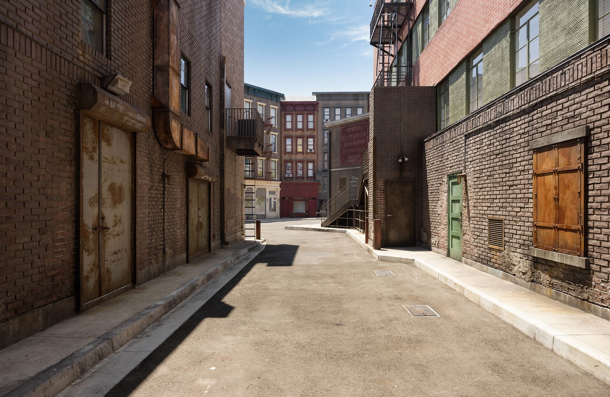 Empty city alleyway between old brick buildings with various doors and a boarded window under a clear blue sky.