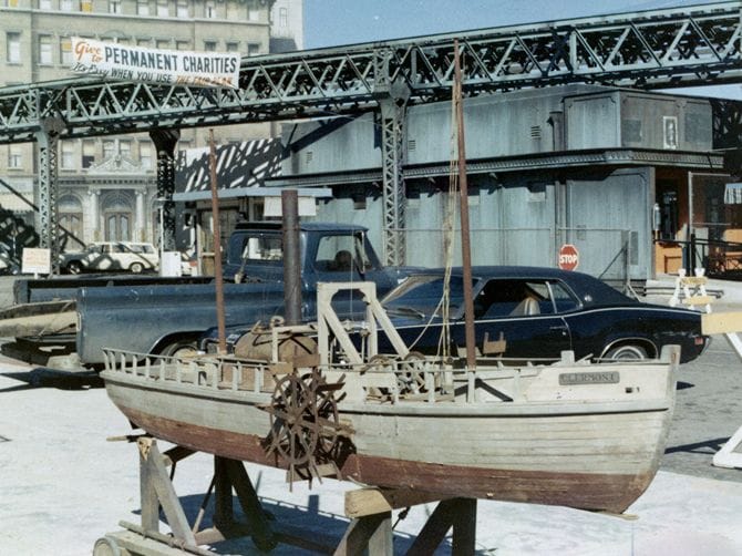 A model of the paddle steamer Clermont on a stand, with cars and city buildings under an elevated train track in the background.