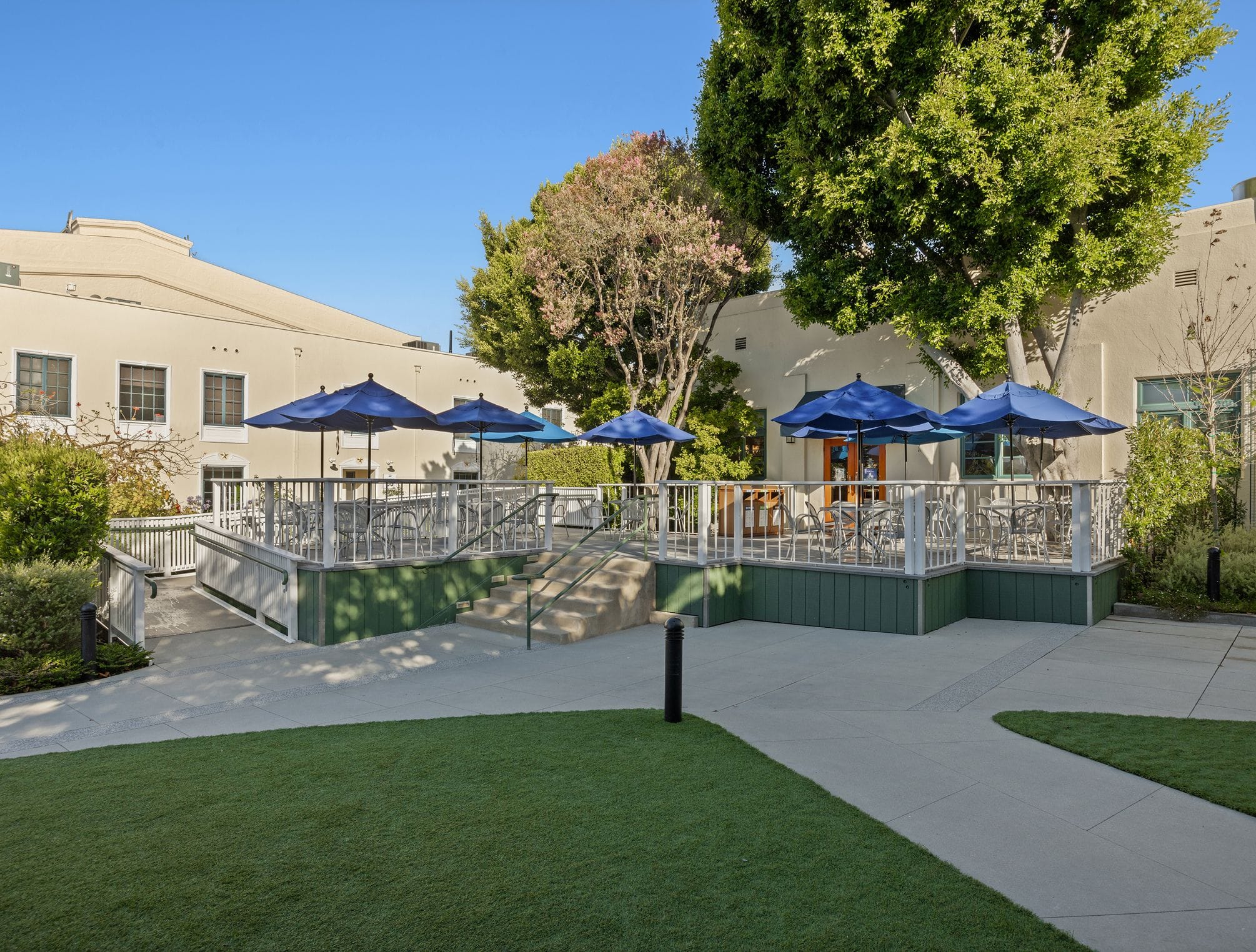 Outdoor patio with blue umbrellas, tables, and chairs, surrounded by buildings and a green lawn.