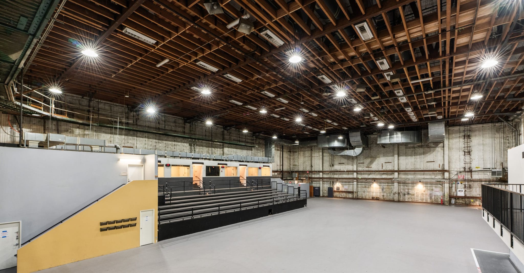 A vast, empty industrial studio or event space with a high wooden beamed ceiling, bright lights, and tiered seating.