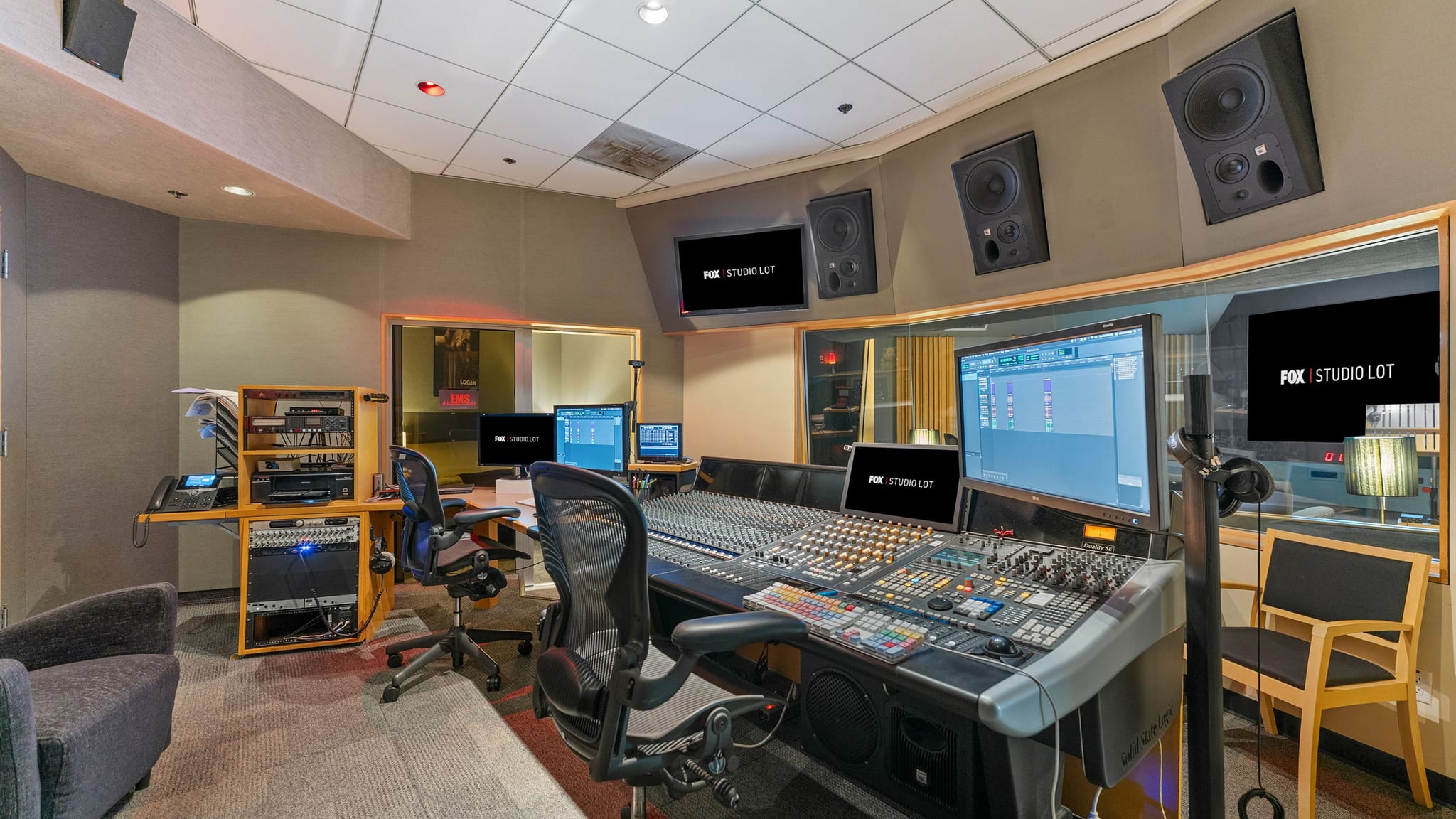 Control room of a sound studio with a large mixing console, multiple monitors displaying "FOX STUDIO LOT", and wall-mounted speakers.