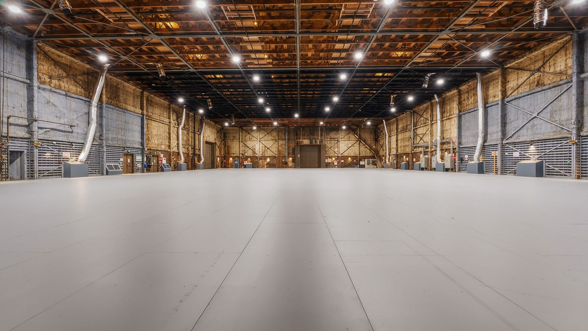 A vast, empty industrial space with a light gray floor, high exposed wooden ceiling, and industrial lighting.