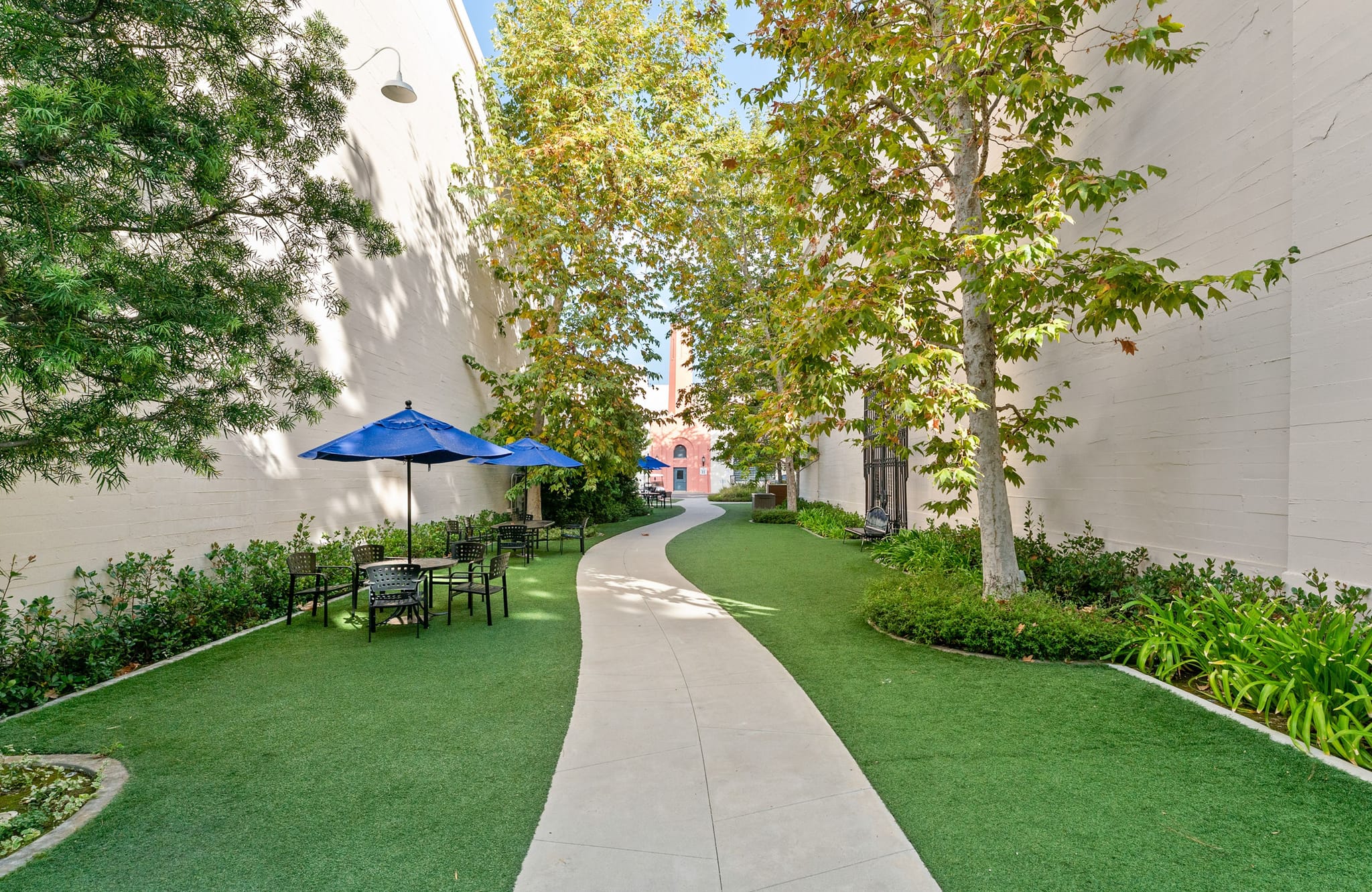 A curved concrete path winds through a green courtyard with trees, white walls, and outdoor seating under blue umbrellas.
