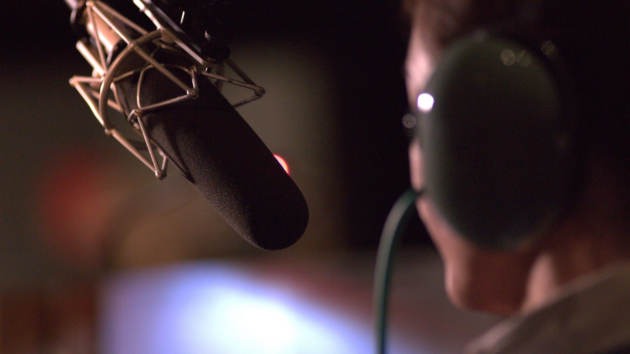 A studio microphone in the foreground with a person wearing headphones in the blurry background.