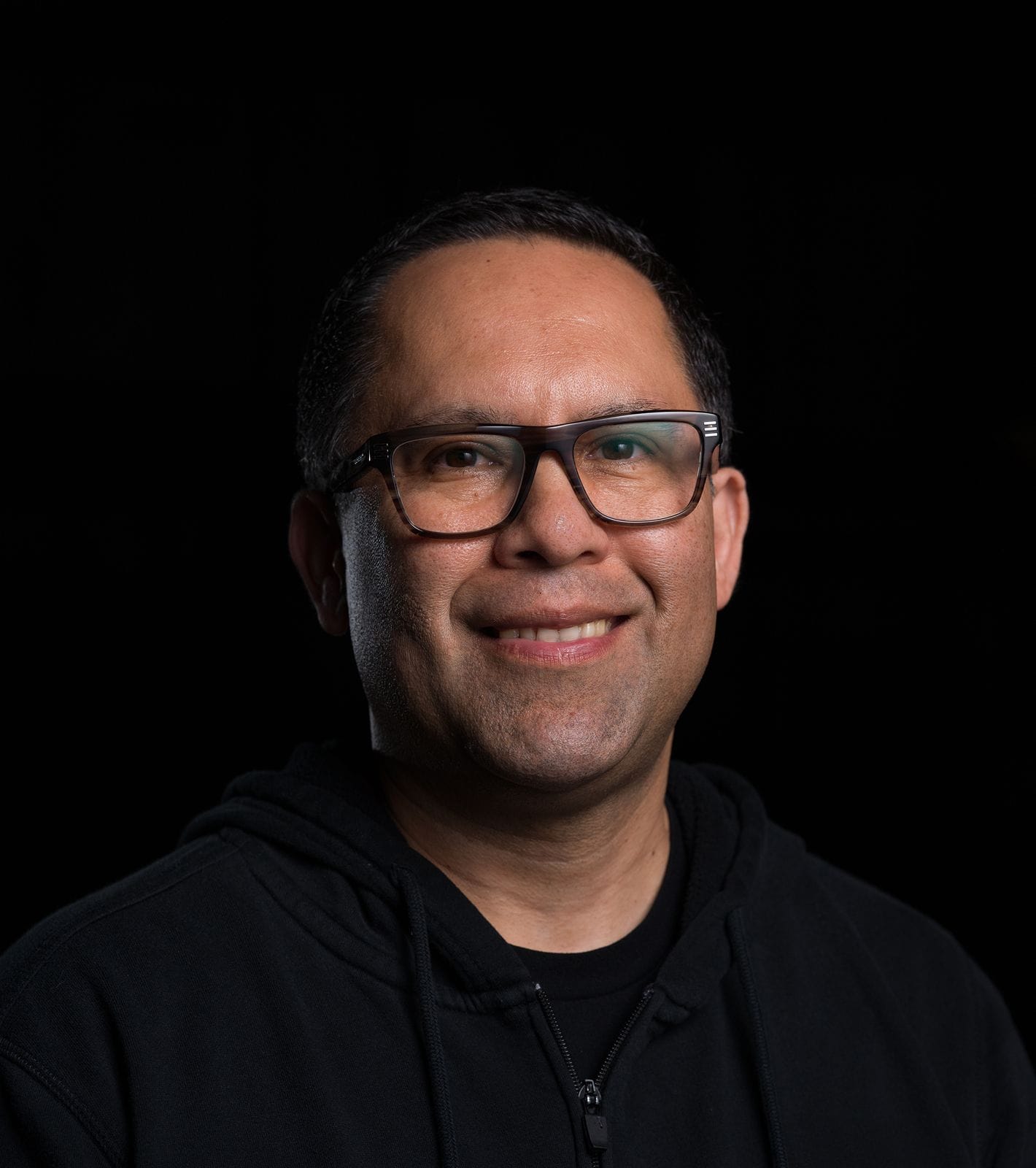 A smiling man with dark hair and glasses wears a black hoodie against a dark background.