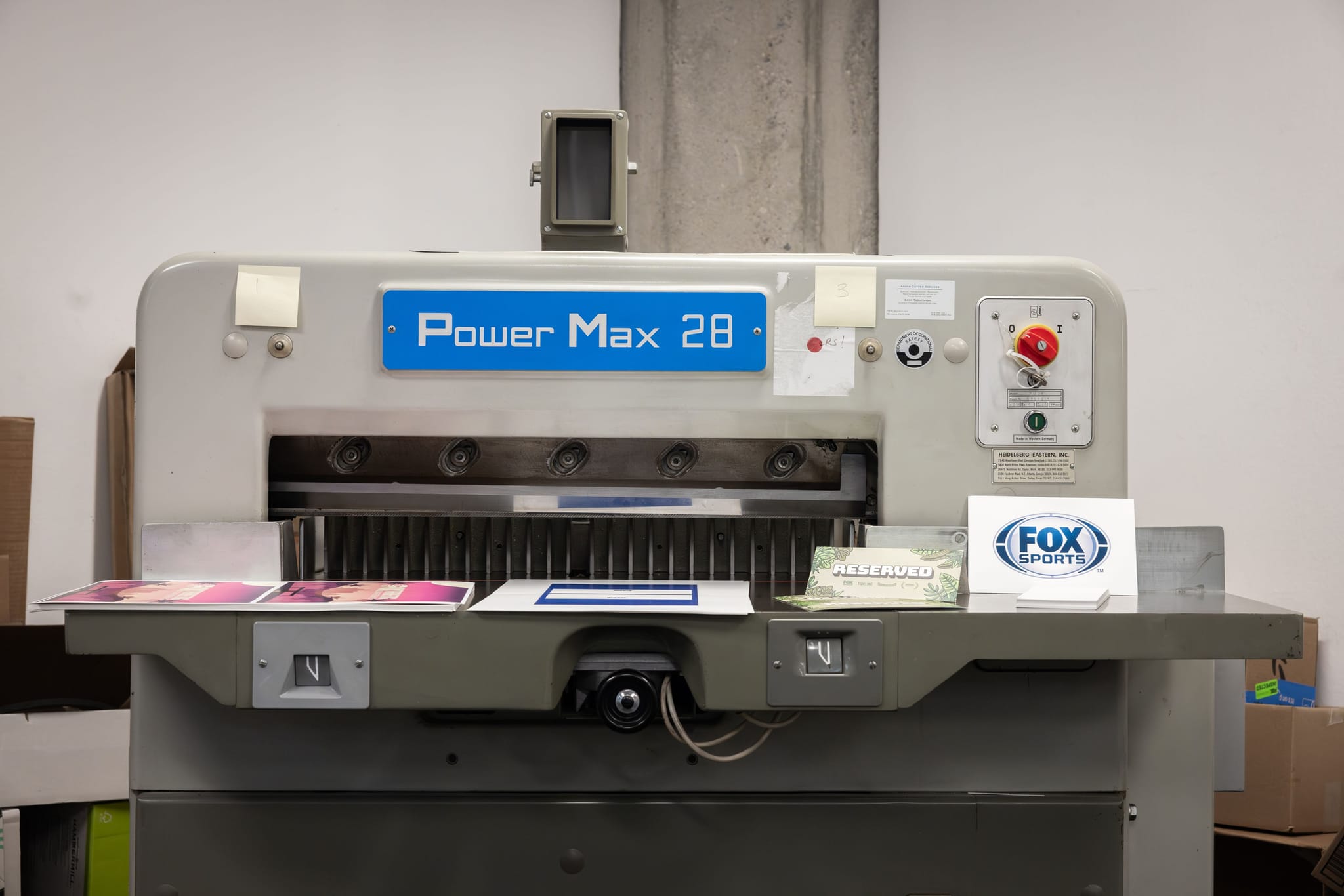 A Power Max 28 industrial paper cutter with papers, including one with a Fox Sports logo, on its tray.